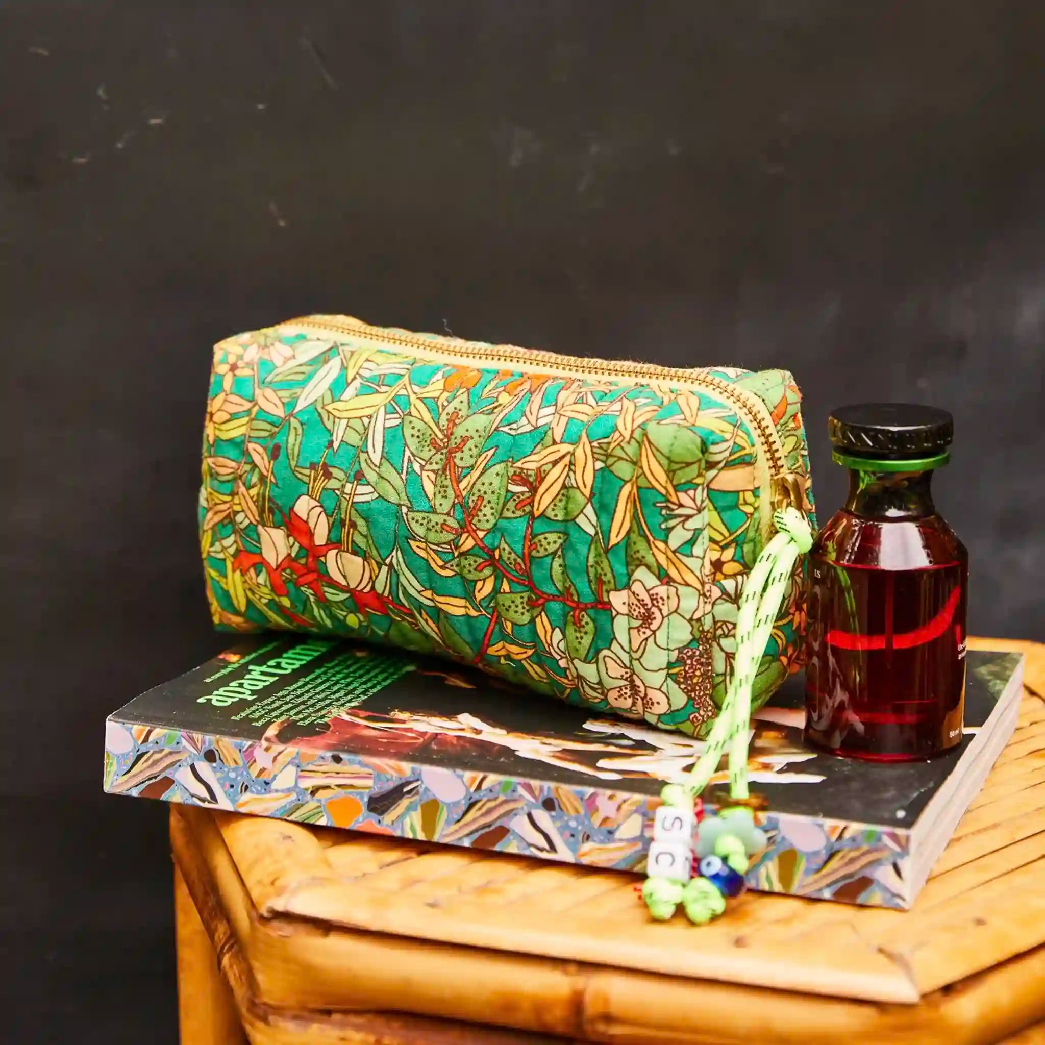 Green floral-patterned pouch on a book with a bottle of liquid and a magazine on a wooden surface.