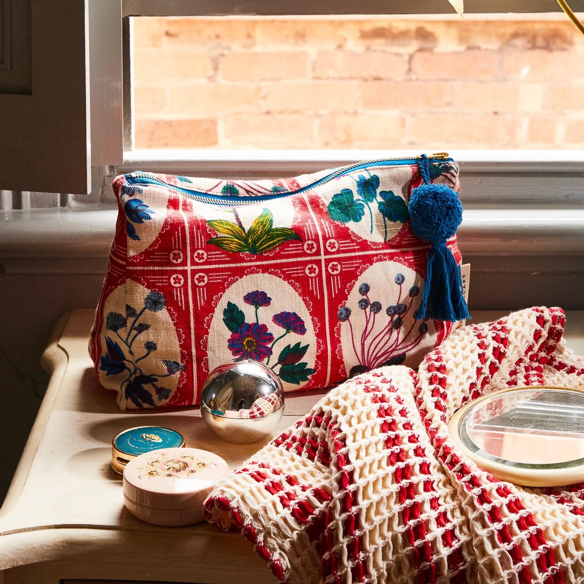 A red and multicolored floral print organic cotton cosmetic bag with a tassel zipper pull in a bathroom.