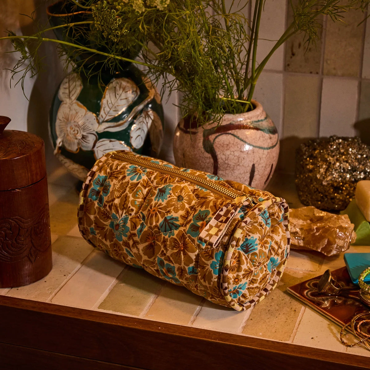 Sage x Clare Magnolia Makeup Bag - Turquoise on a tiled bathroom shelf beside ceramic pots and plants