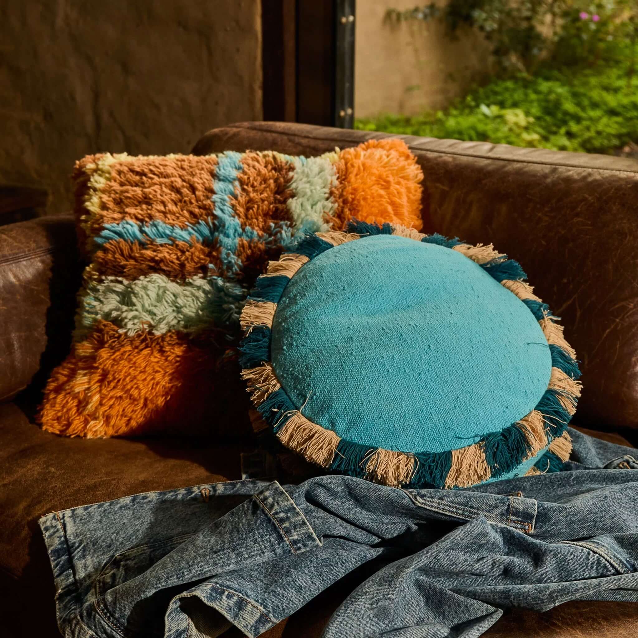Sage x Clare Reese Round Cushion - Turqouise on a leather sofa with a colorful fluffy cushion.