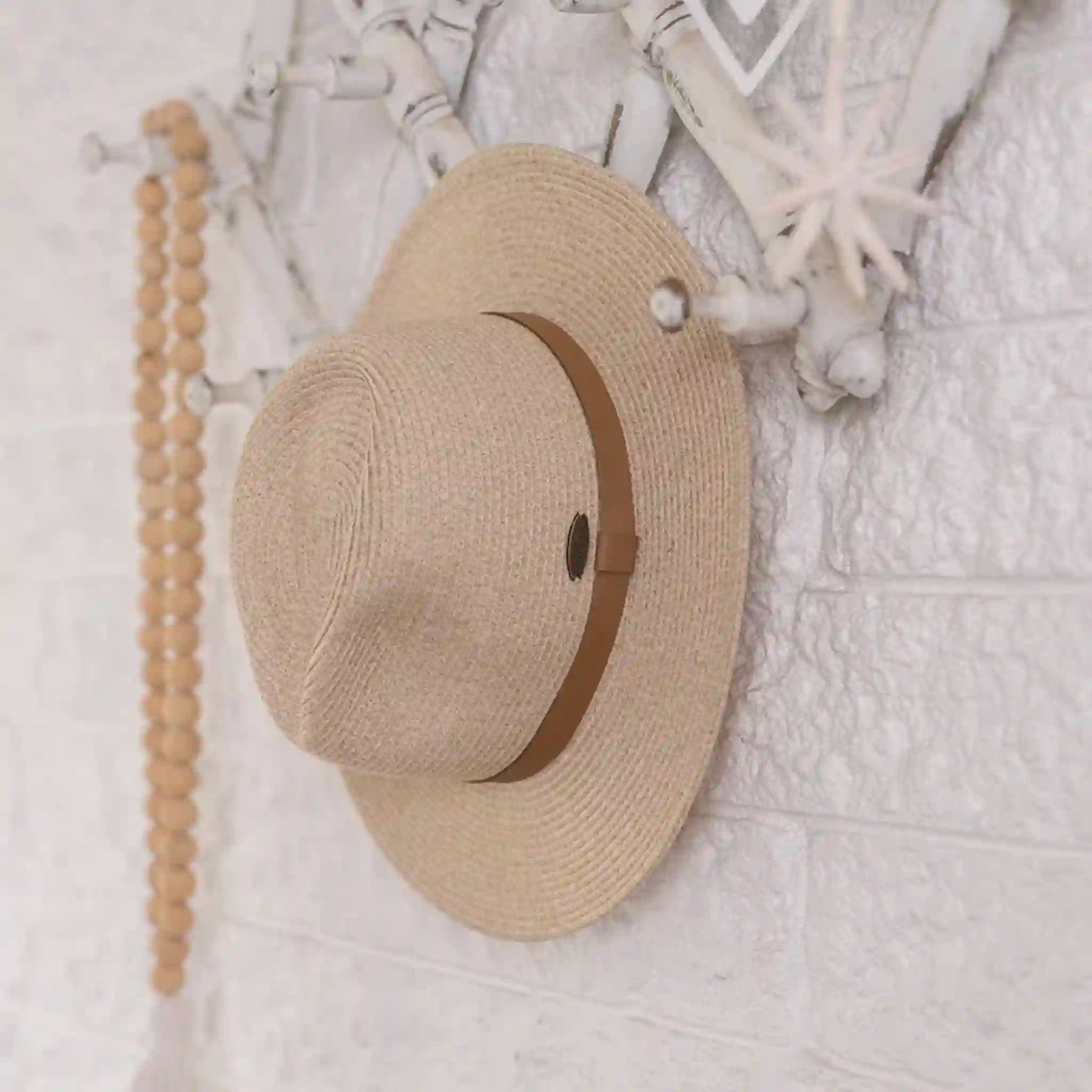 Beige straw hat with a brown band hanging on a white textured wall.