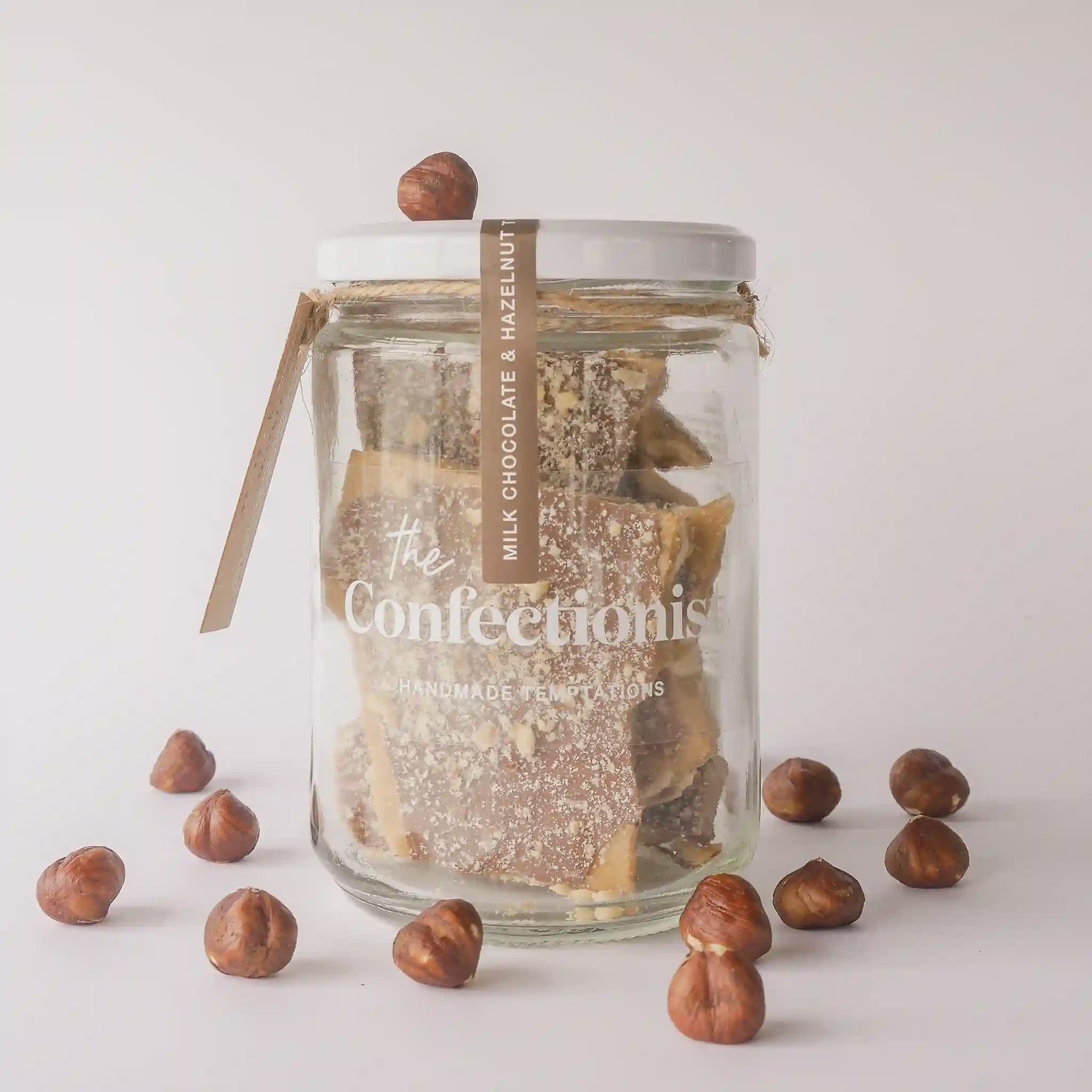 Glass jar filled with chocolate bars labeled 'The Confectionist' surrounded by hazelnuts on a light background