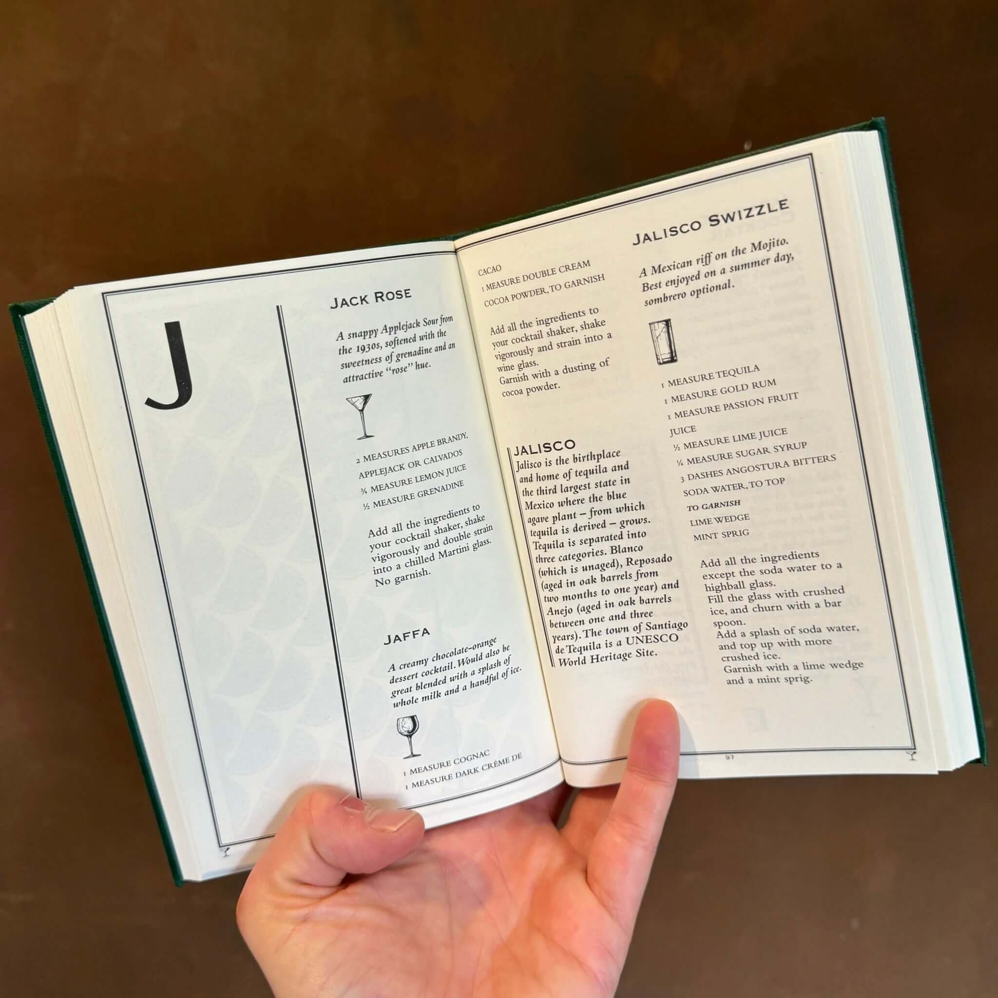 A person holding open The Cocktail Bible displaying cocktail recipes for Jack Rose and Jalisco Swizzle.