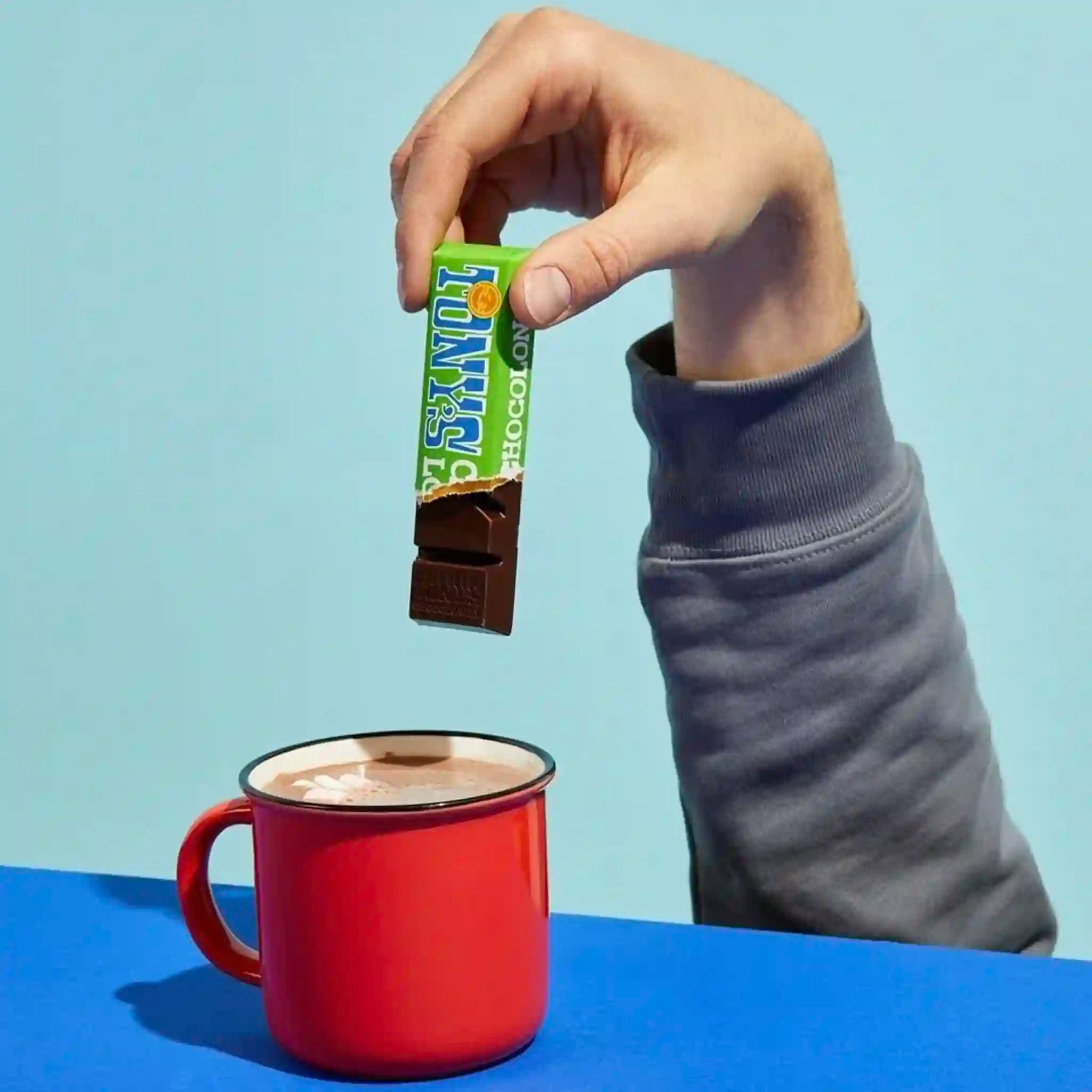 Hand holding a chocolate bar above a red mug on a blue background