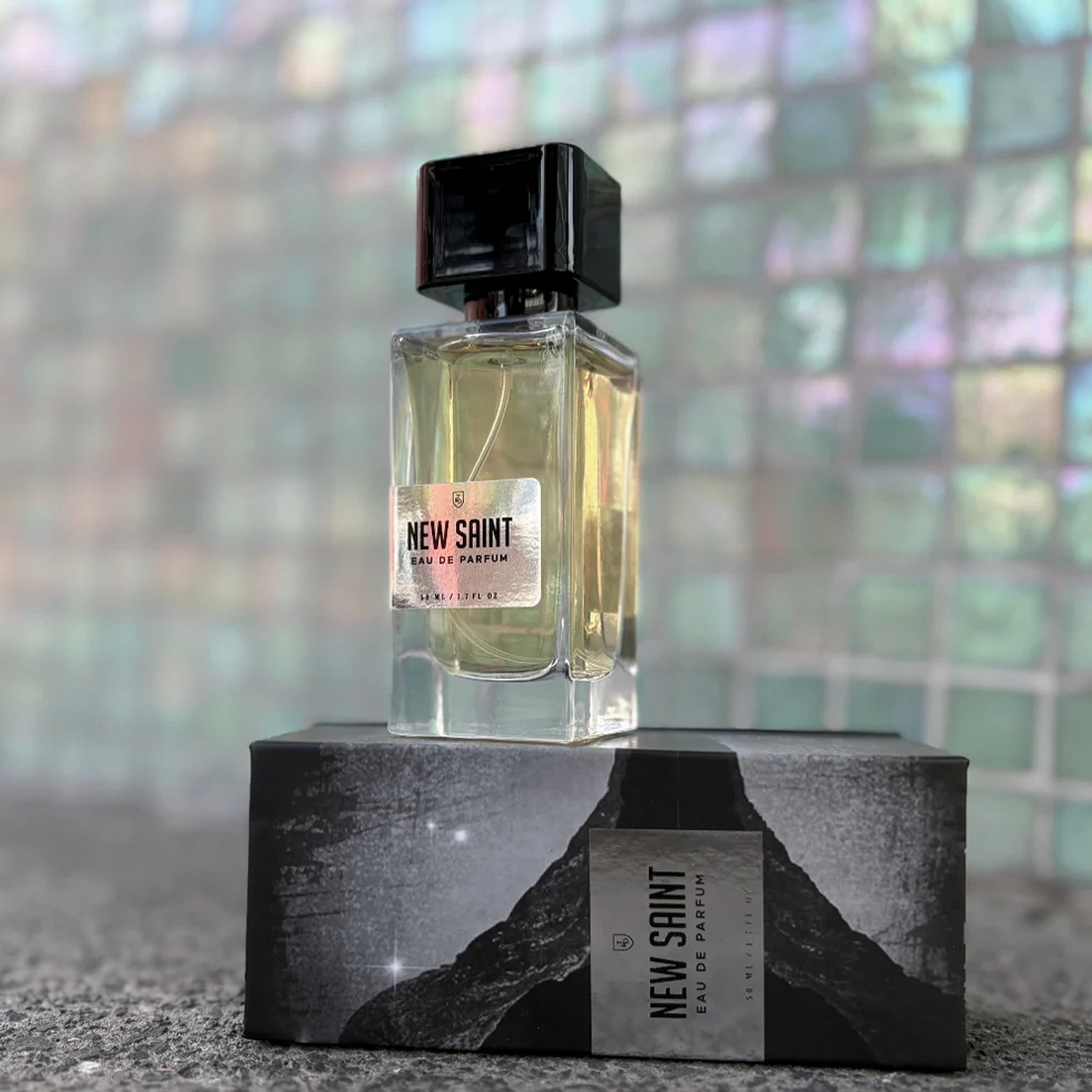 Perfume bottle labeled 'New Saint' on a black stand with a blurred multicolored background