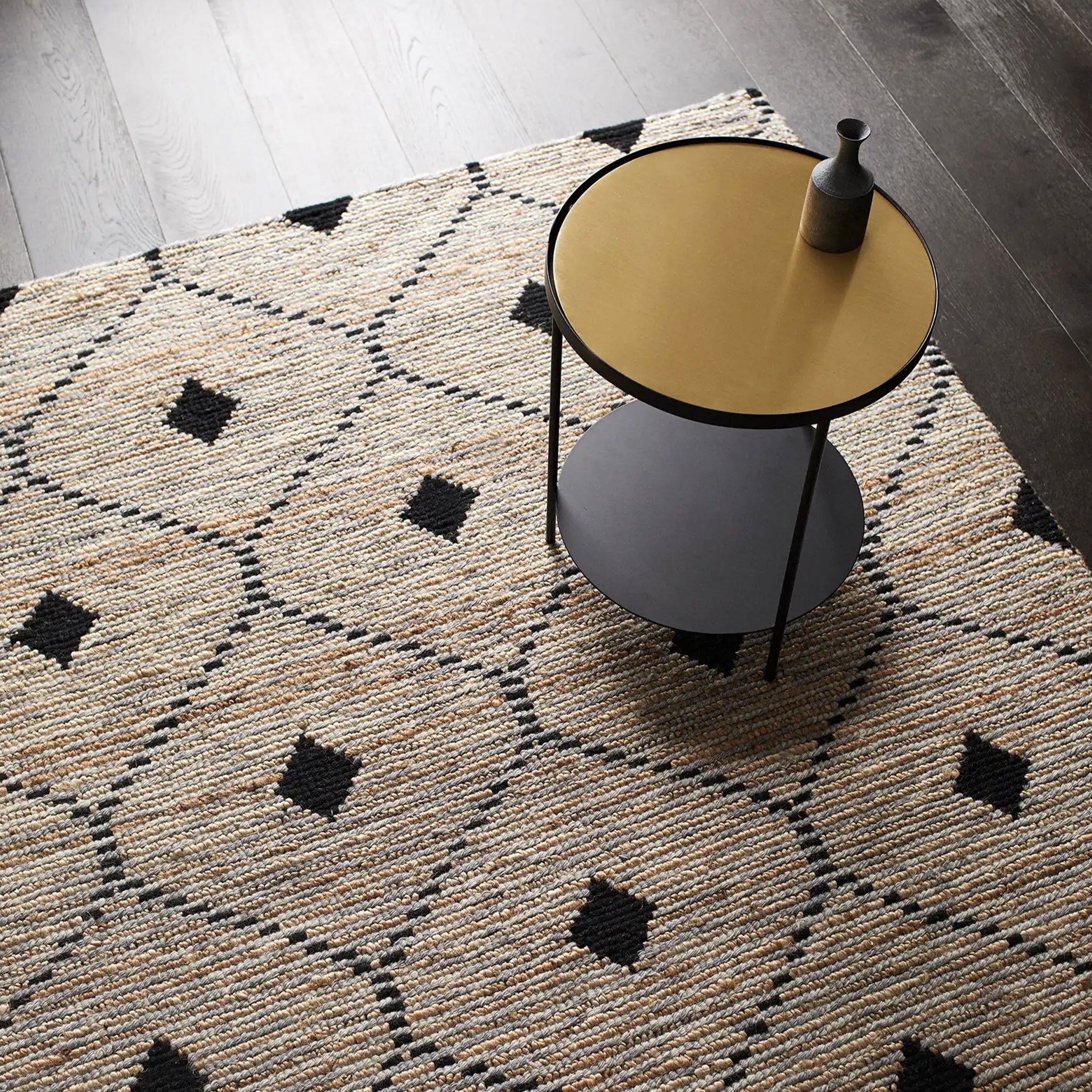 A rectangular, flatweave rug with a geometric pattern in beige, black, and white colors, featuring a round table and lamp on top of it.