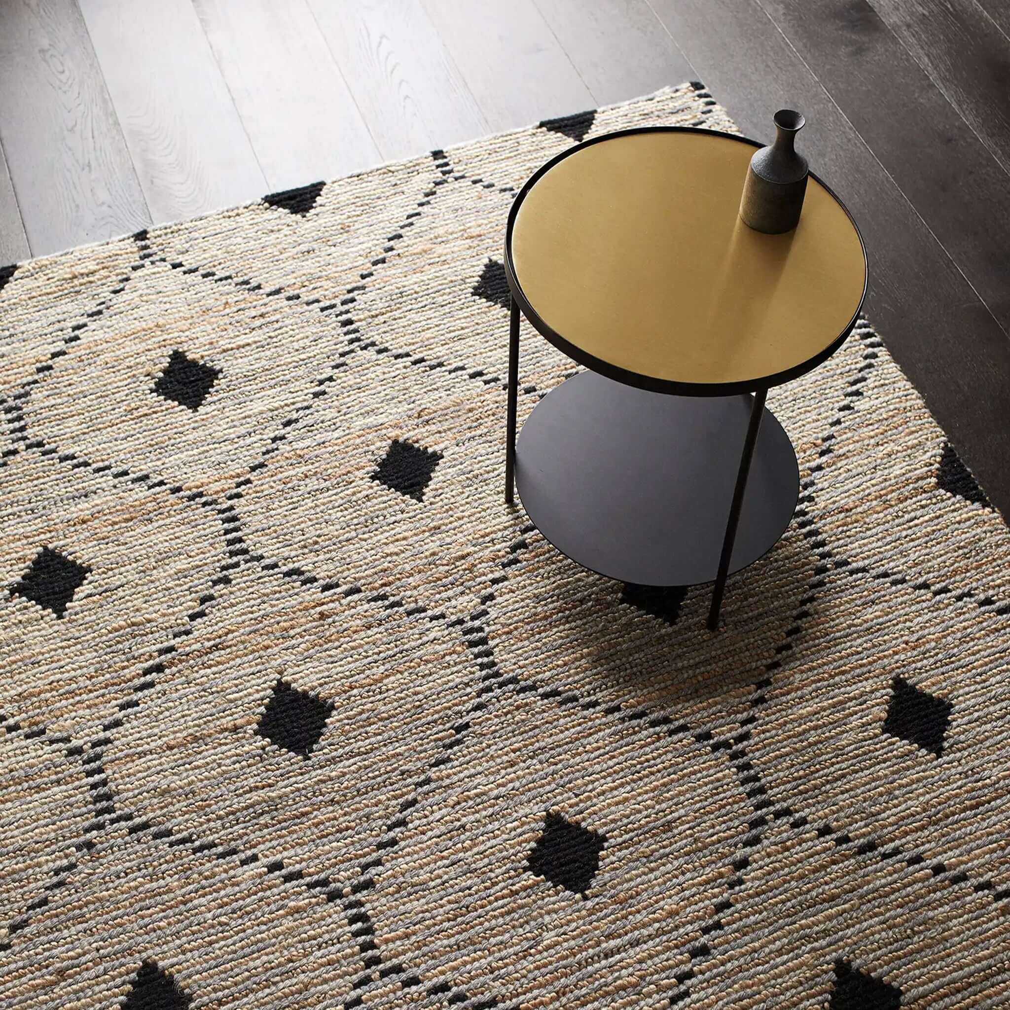 A rectangular, flatweave rug with a geometric pattern in beige, black, and white colors, featuring a round table and lamp on top of it.