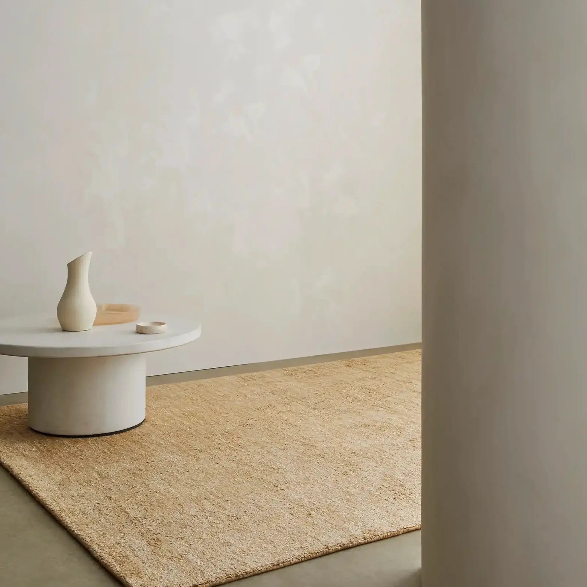 Beige rug on a neutral-colored floor with a white wall and a small round table with a vase.