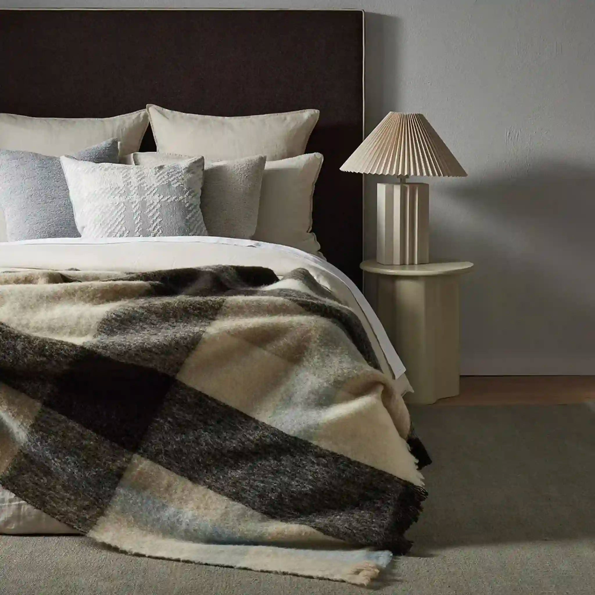 Bed with plaid blanket and pillows next to a lamp on a nightstand.