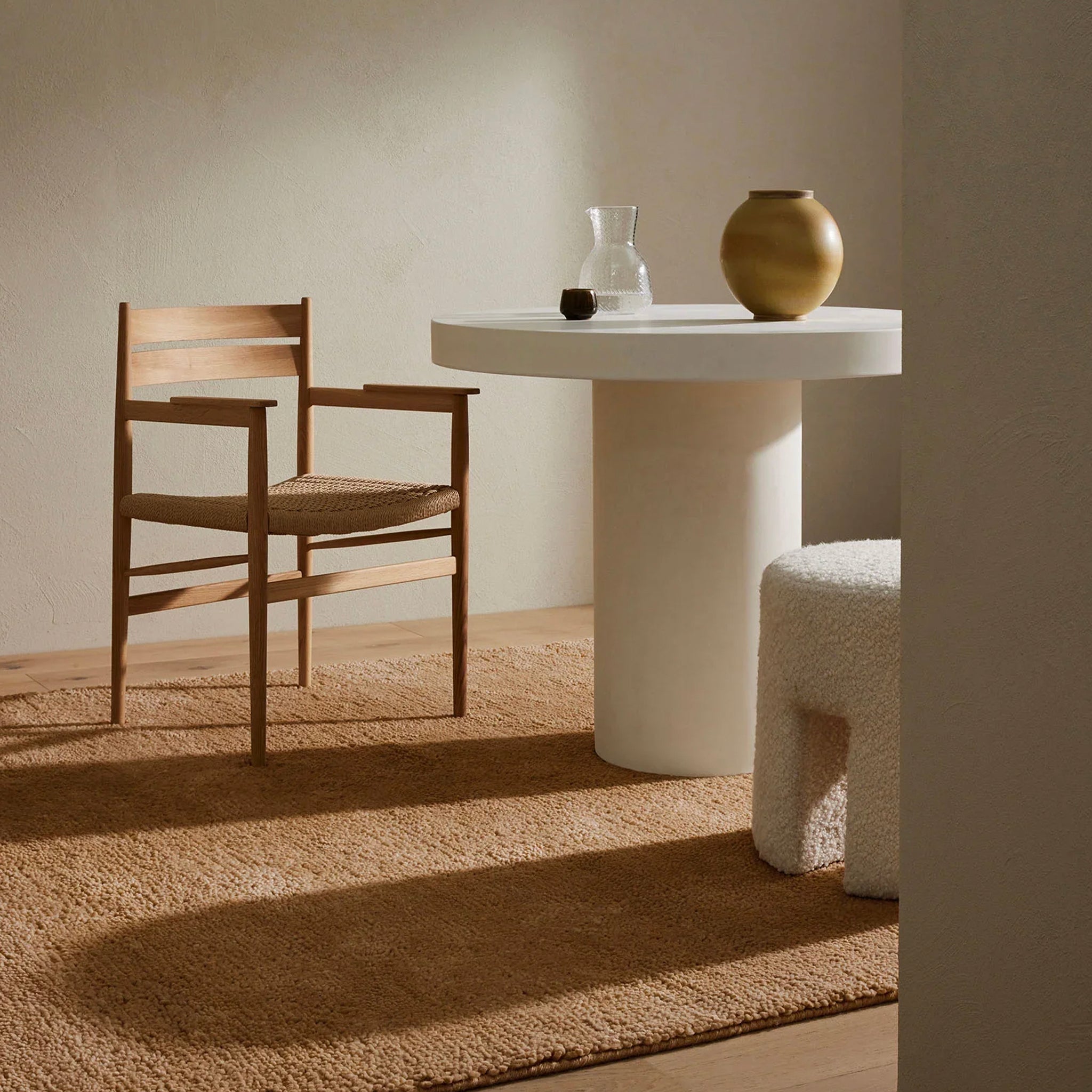 A well-lit image showing a beige wool rug under a white table with a vase and a gold-colored sculpture, with a wooden chair to the side in a cozy indoor setting.
