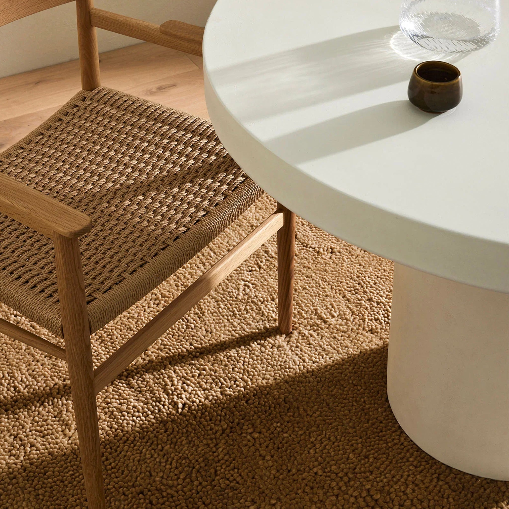 A plush, chunky woven style rug in a brown color, made with 100% wool pile, displayed under a round white table with a glass vase and a dark object on top.