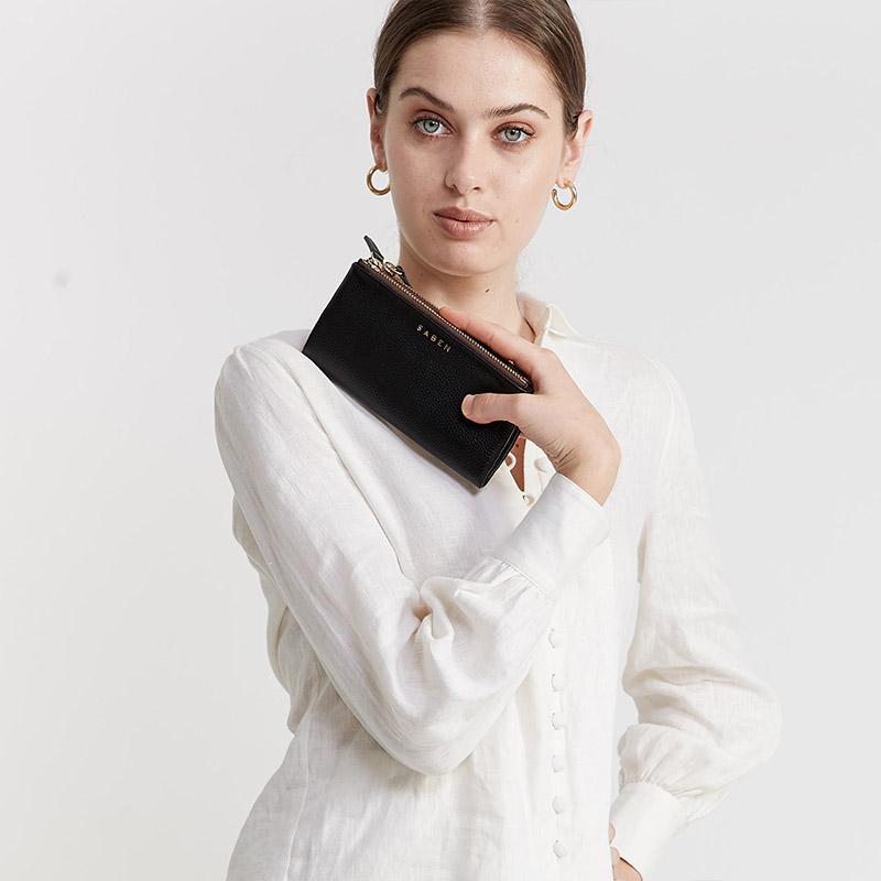 Woman holding a black leather wallet with gold zipper and 'SABEN' branding on a white background