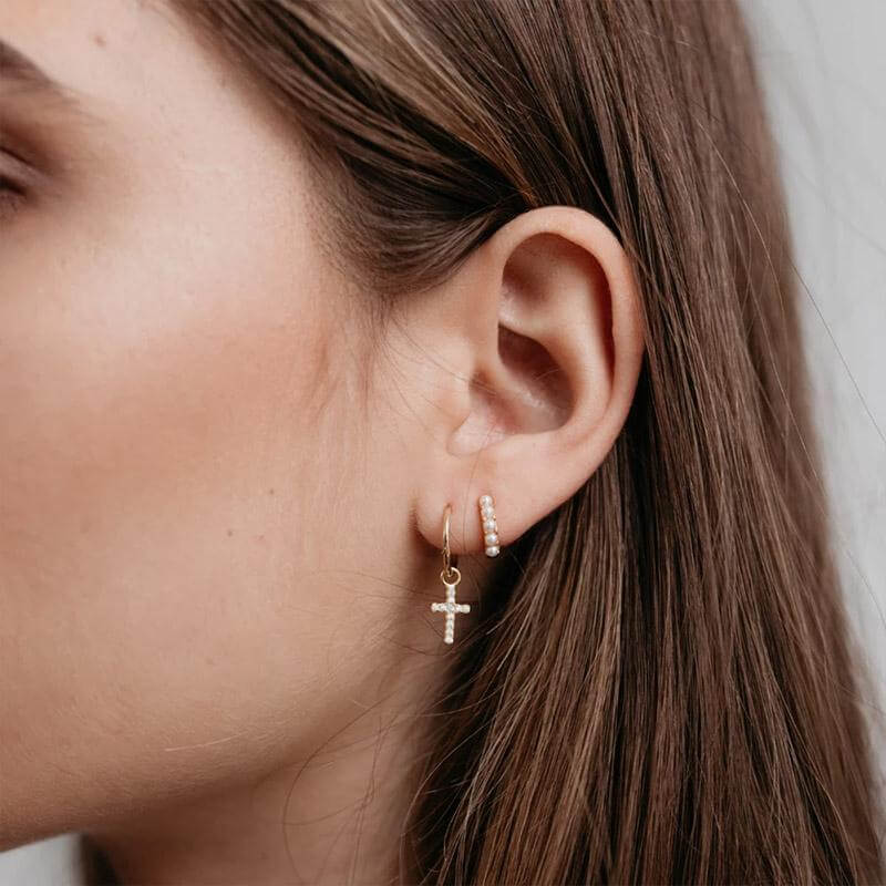 Alt Tag: Sophie Mini Pearl Huggie Earrings worn on ear with mini faux pearl huggie hoop and cross charm, delicate layered earring look