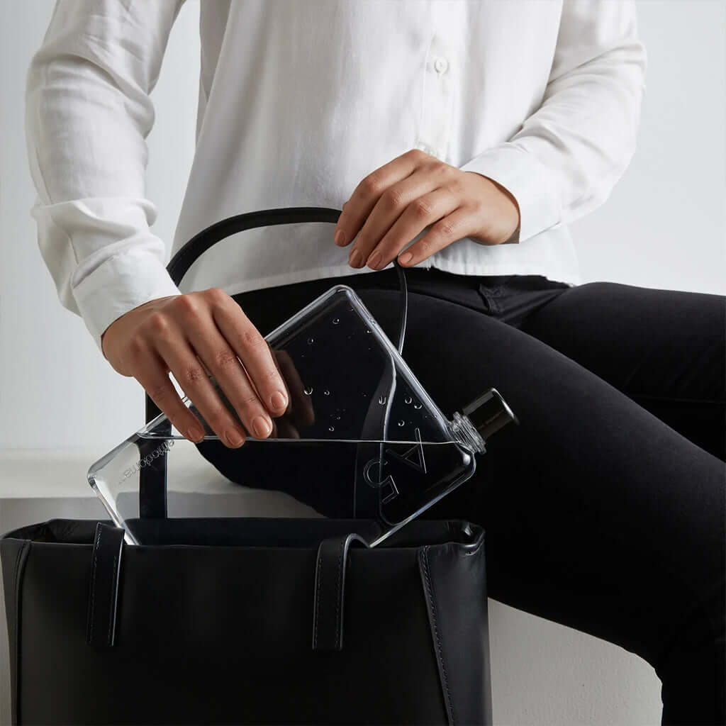 Memobottle A5 - Transparent flat water bottle sliding into a tote bag beside a tablet, minimalist reusable hydration for work or travel