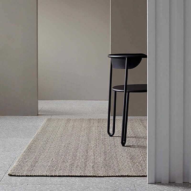 A rectangular, flatweave rug with a braided edge and a textured surface, primarily in a neutral color palette, placed on a tiled floor next to a black chair.
