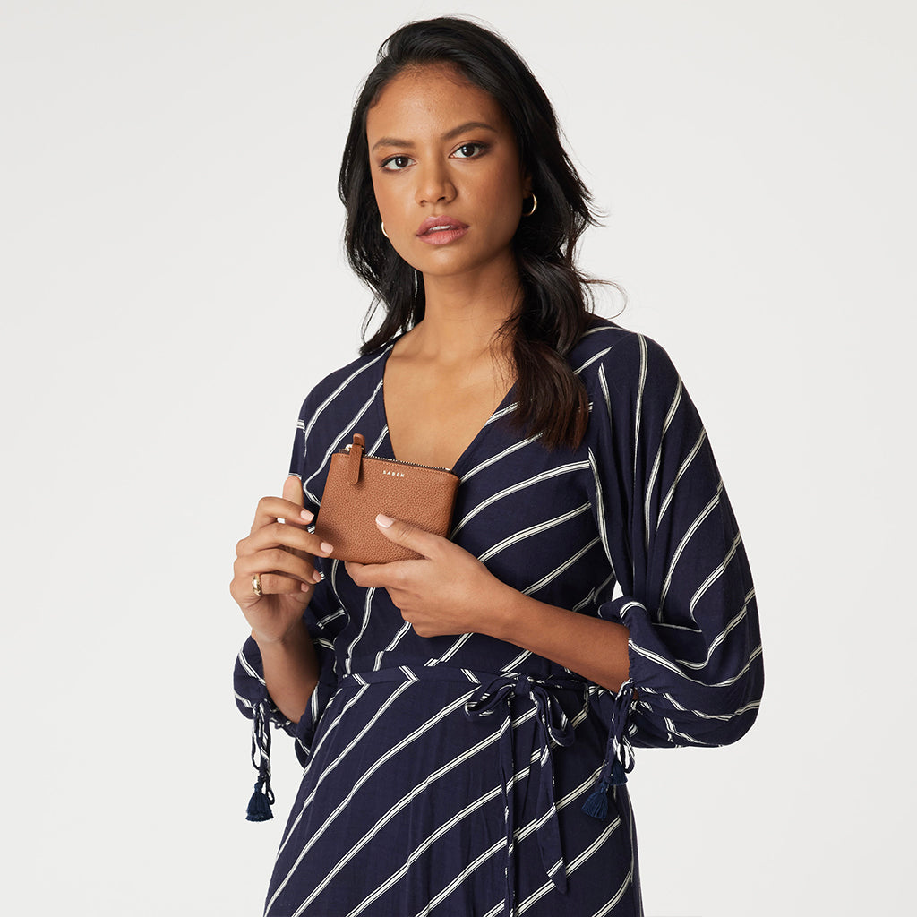 Woman holding a brown leather wallet with gold zipper and 'SABEN' branding on a neutral background