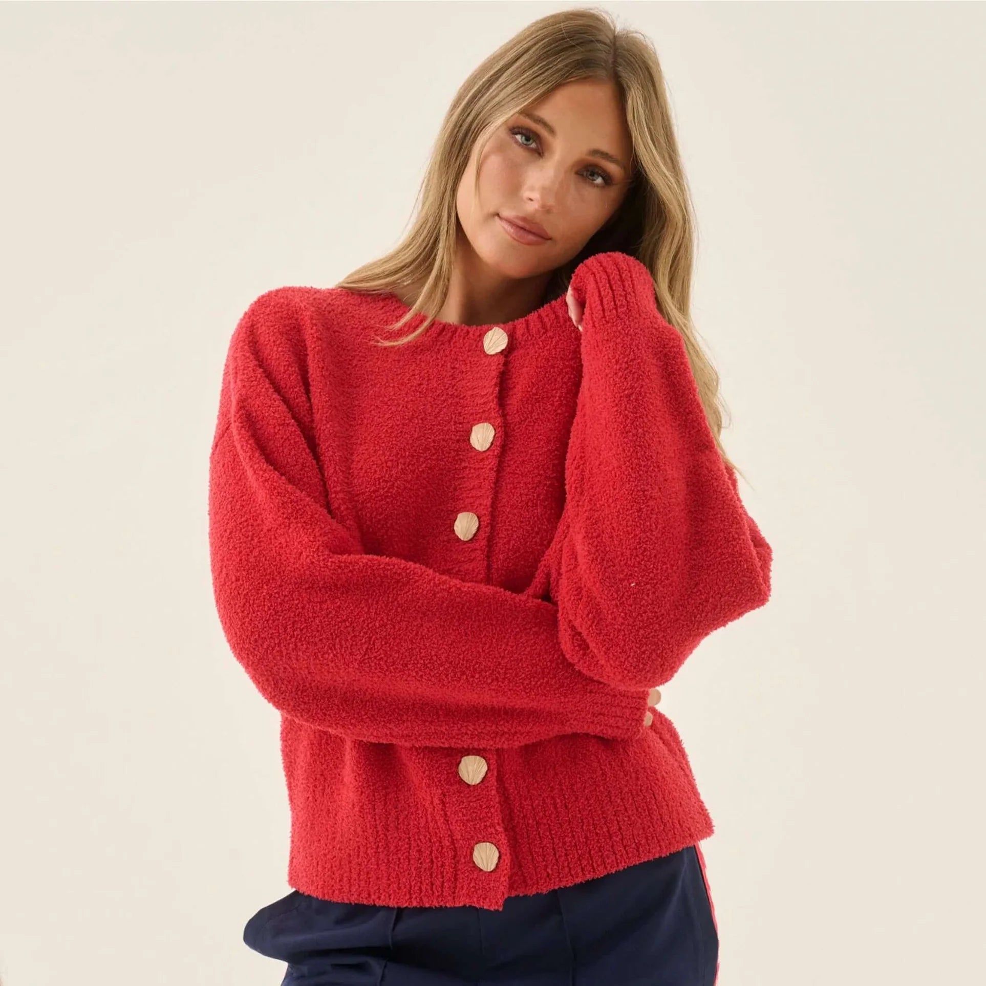 A woman wearing a bright red fuzzy knit cardigan with cream buttons, posed against a plain light beige studio background.