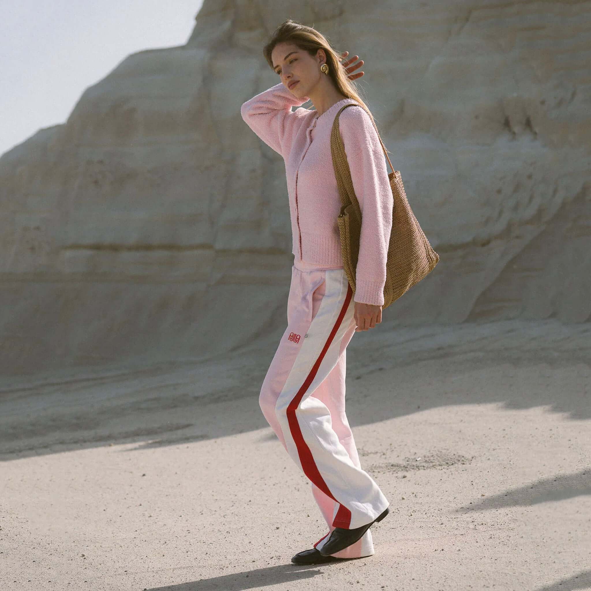 Woman wearing Araminta James Mallorca Trackpants - Ballet Pink with a pink cardigan and tote bag on a sandy background.