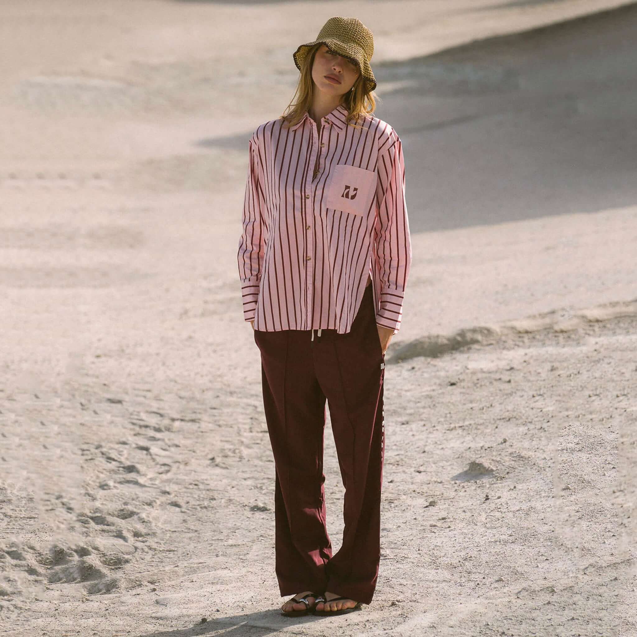 Model wearing Araminta James Pomodoro Twill Pants - Burgundy with a striped shirt and hat in a sandy landscape.