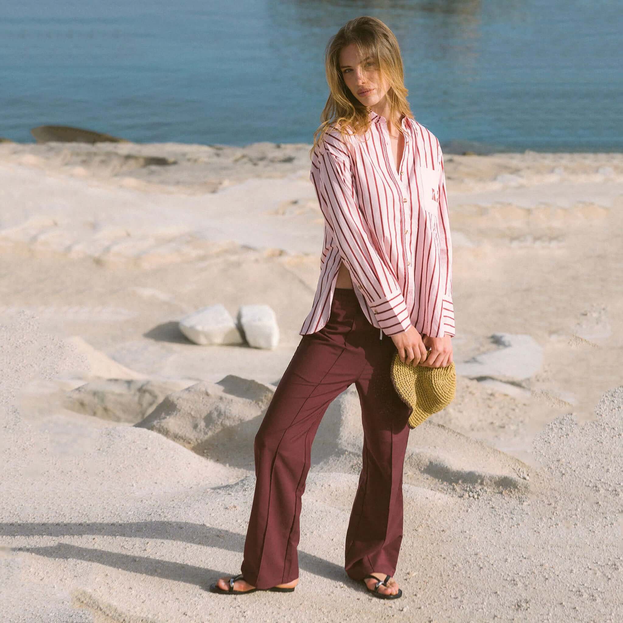 Araminta James Pomodoro Twill Pants - Burgundy worn with a striped shirt on a beach.