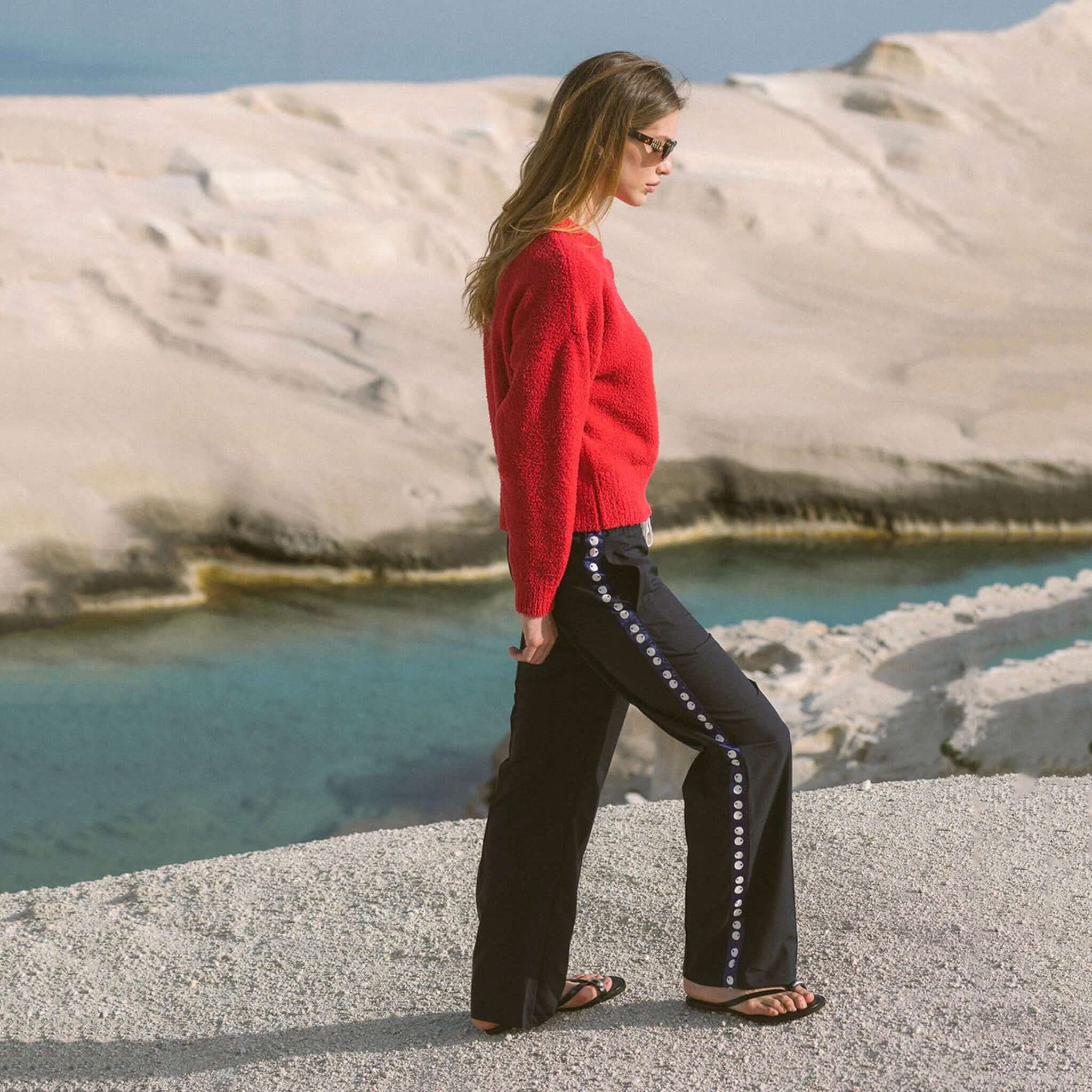 Woman wearing Araminta James Pomodoro Twill Pants - Midnight Navy with red sweater by the sea.
