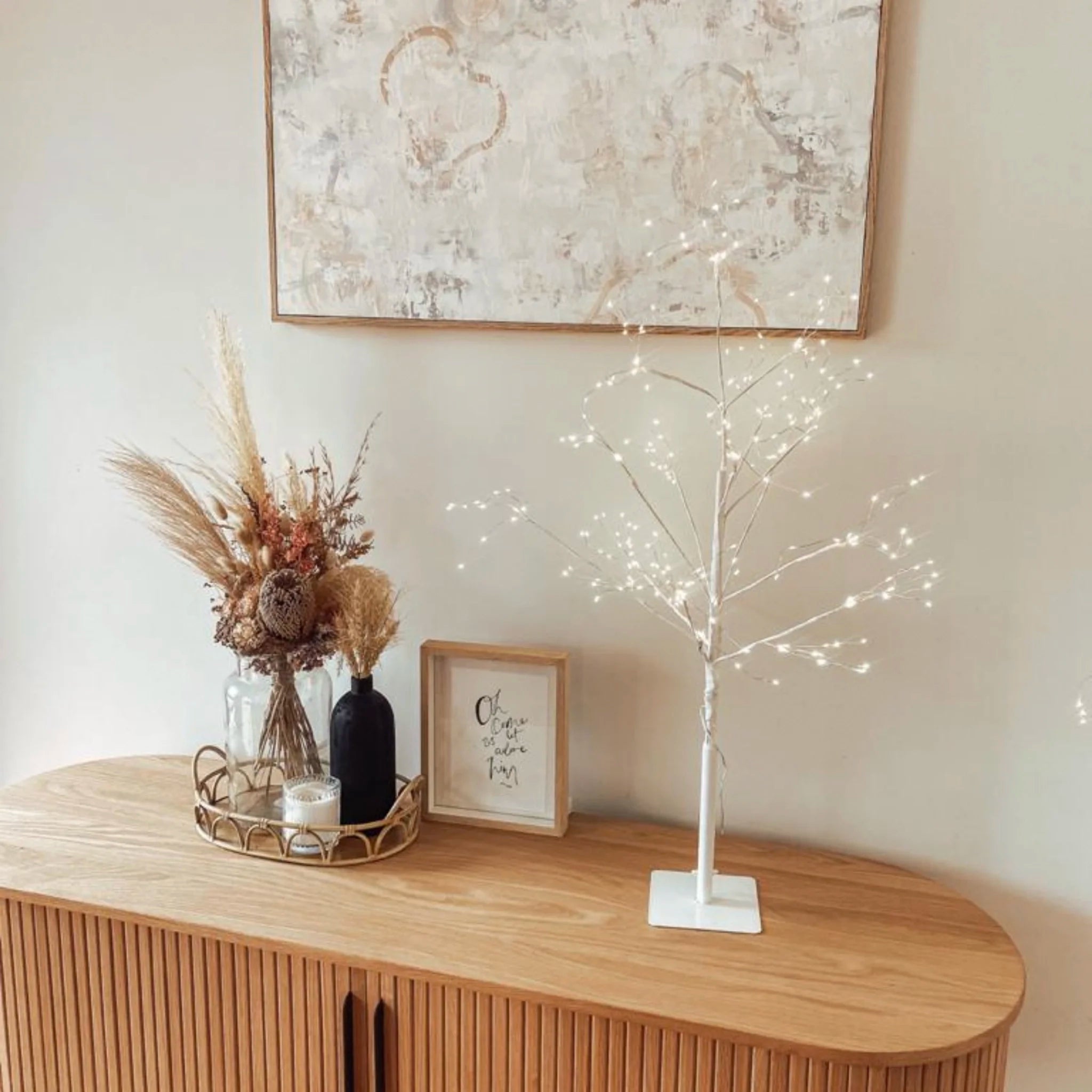 Decorative setup on a wooden console table with a lighted branch, vase with dried flowers, and framed picture.