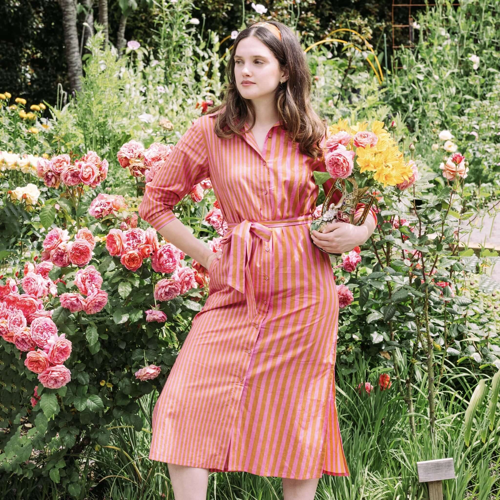 Model wearing the Art In My Wardrobe Clove Dress - Pink surrounded by vibrant roses in a garden.