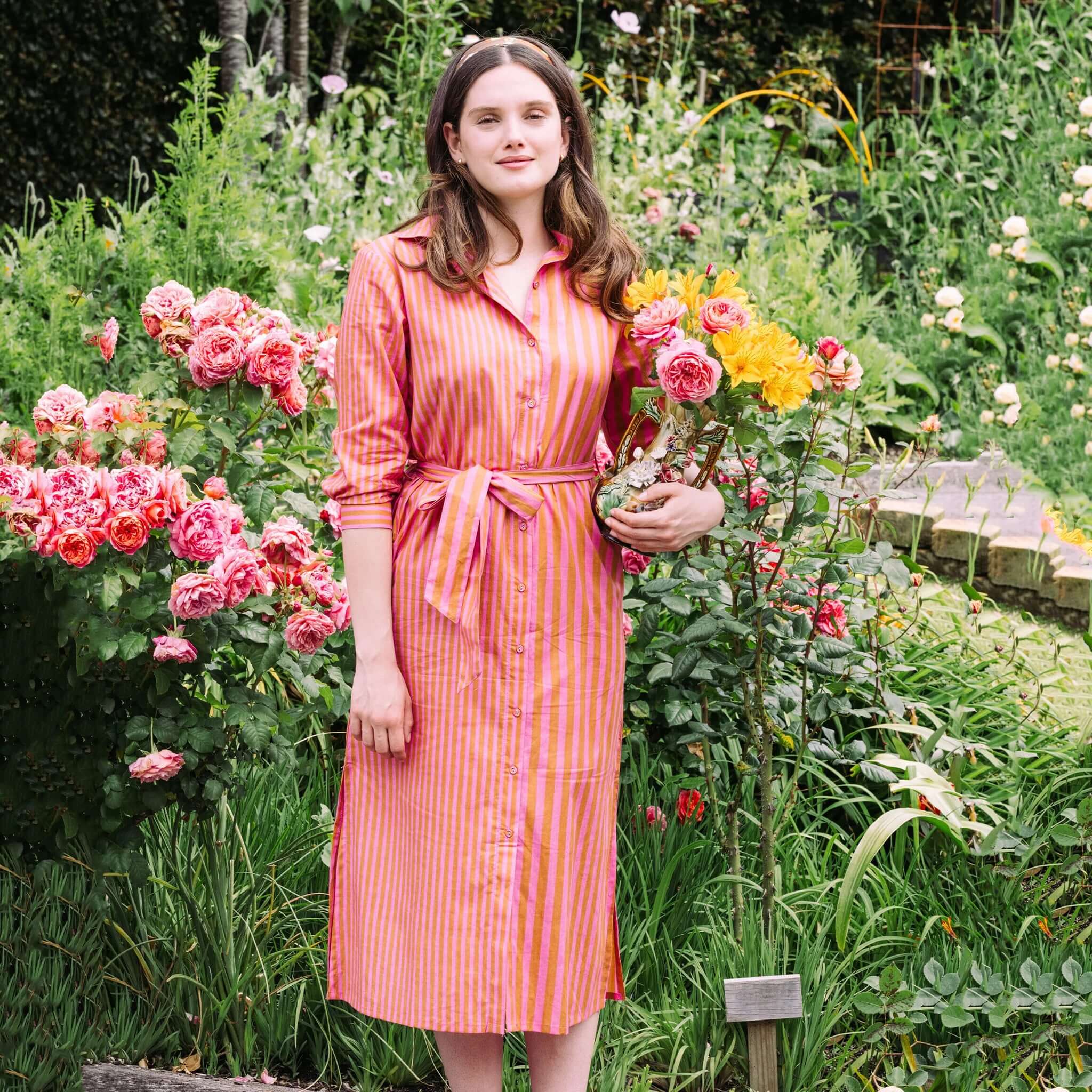 Model wearing the Art In My Wardrobe Clove Dress - Pink in a garden, holding flowers among vibrant roses.