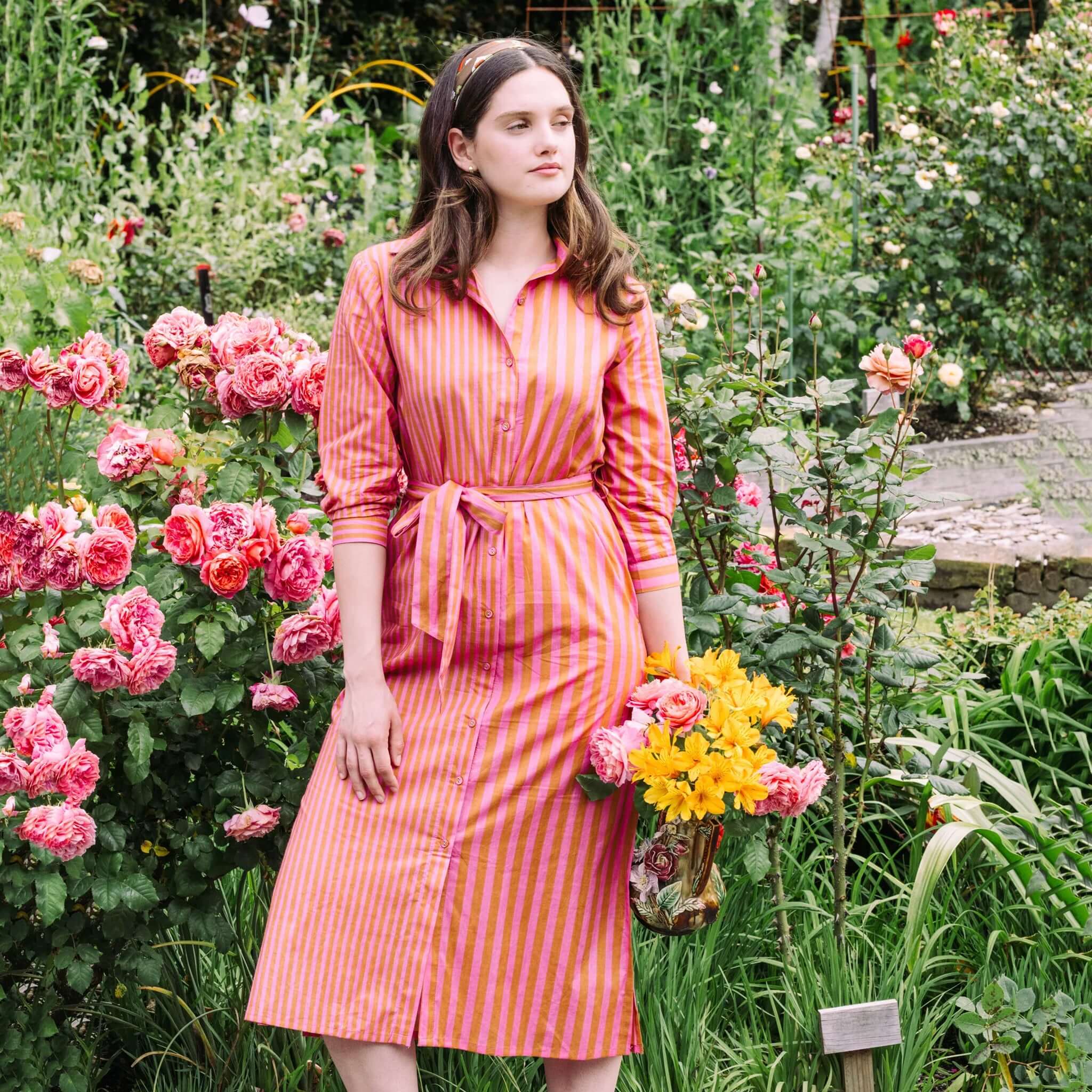 Art In My Wardrobe Clove Dress - Pink worn by a model in a floral garden, showcasing its vibrant stripes and self-tie belt.