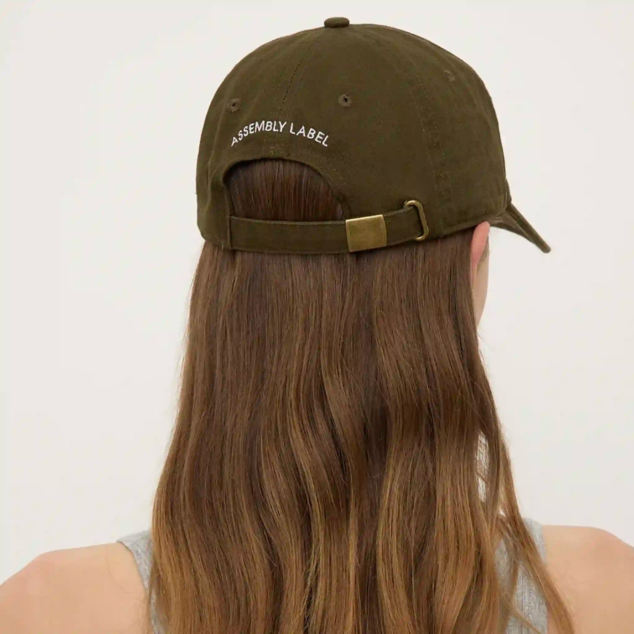 Person wearing an olive green cap with 'Assembly Label' on a white background