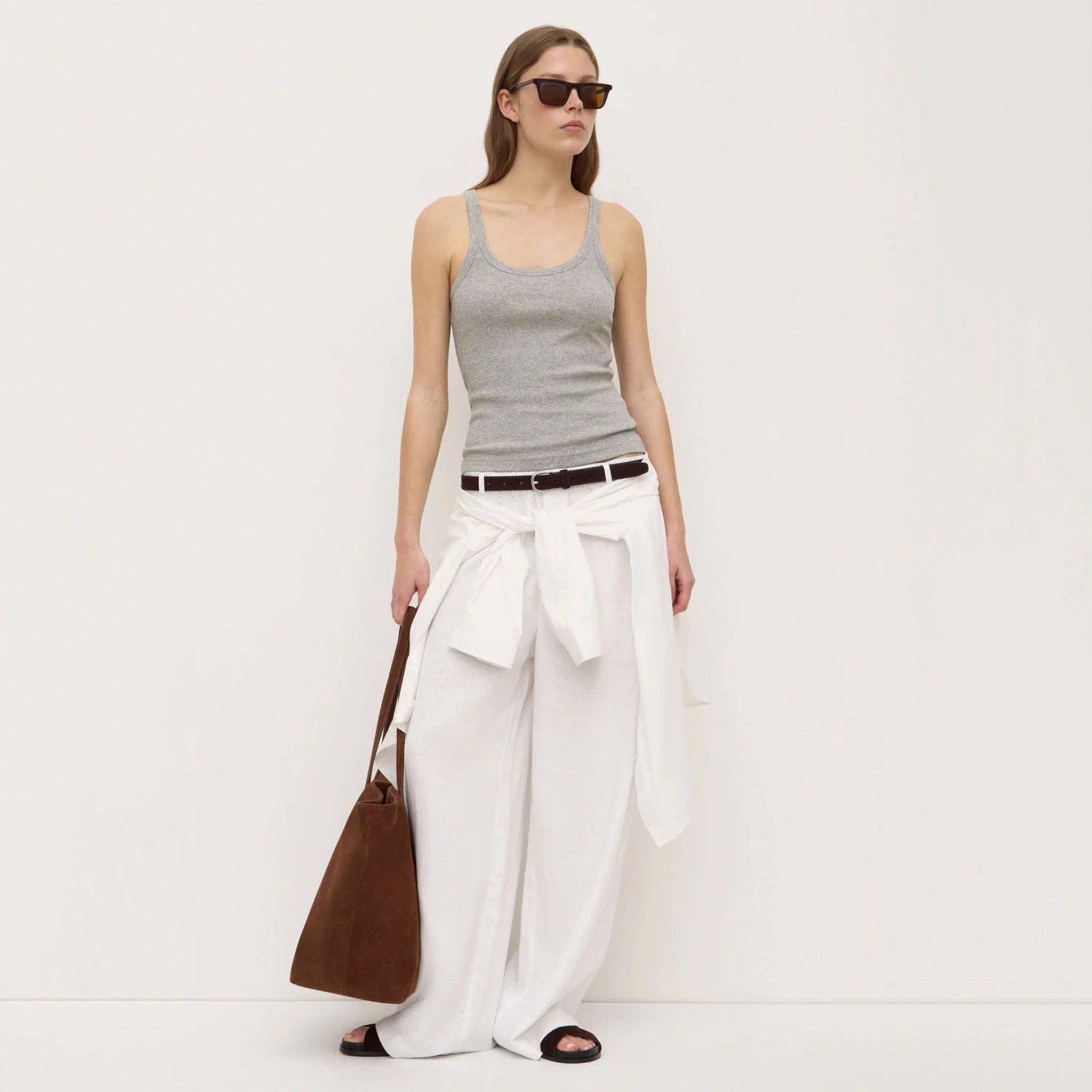 Woman wearing a gray tank top and white wide-leg pants, holding a brown bag on a white background