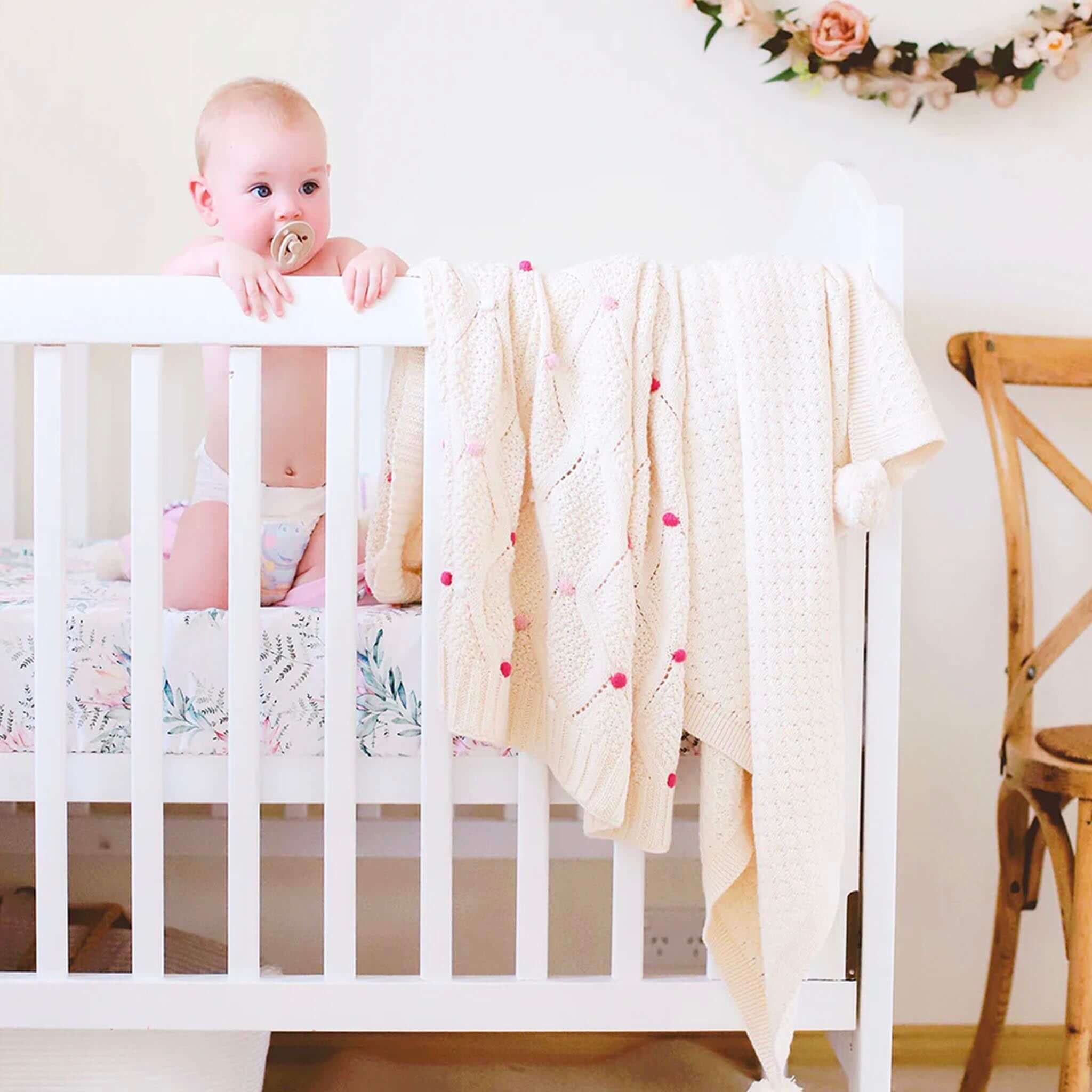 Baby Cotton Lucy Blanket - Ivory & Pink draped on a crib, with a baby looking curiously over the edge.