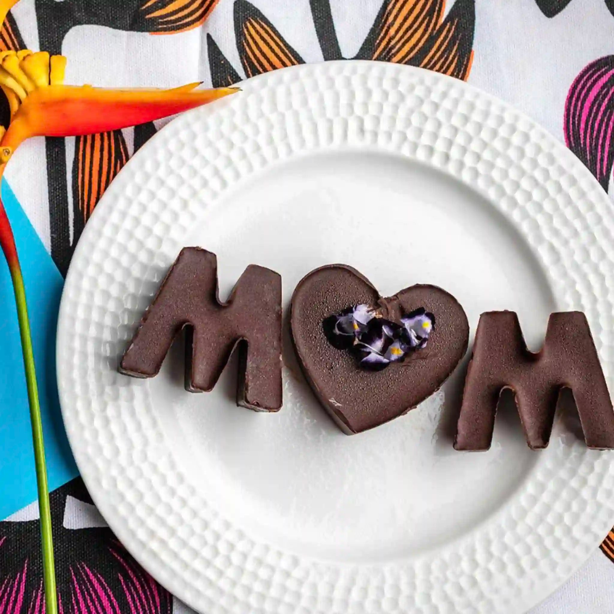 Chocolate dessert shaped like the word 'MOM' on a white plate with a colorful background.
