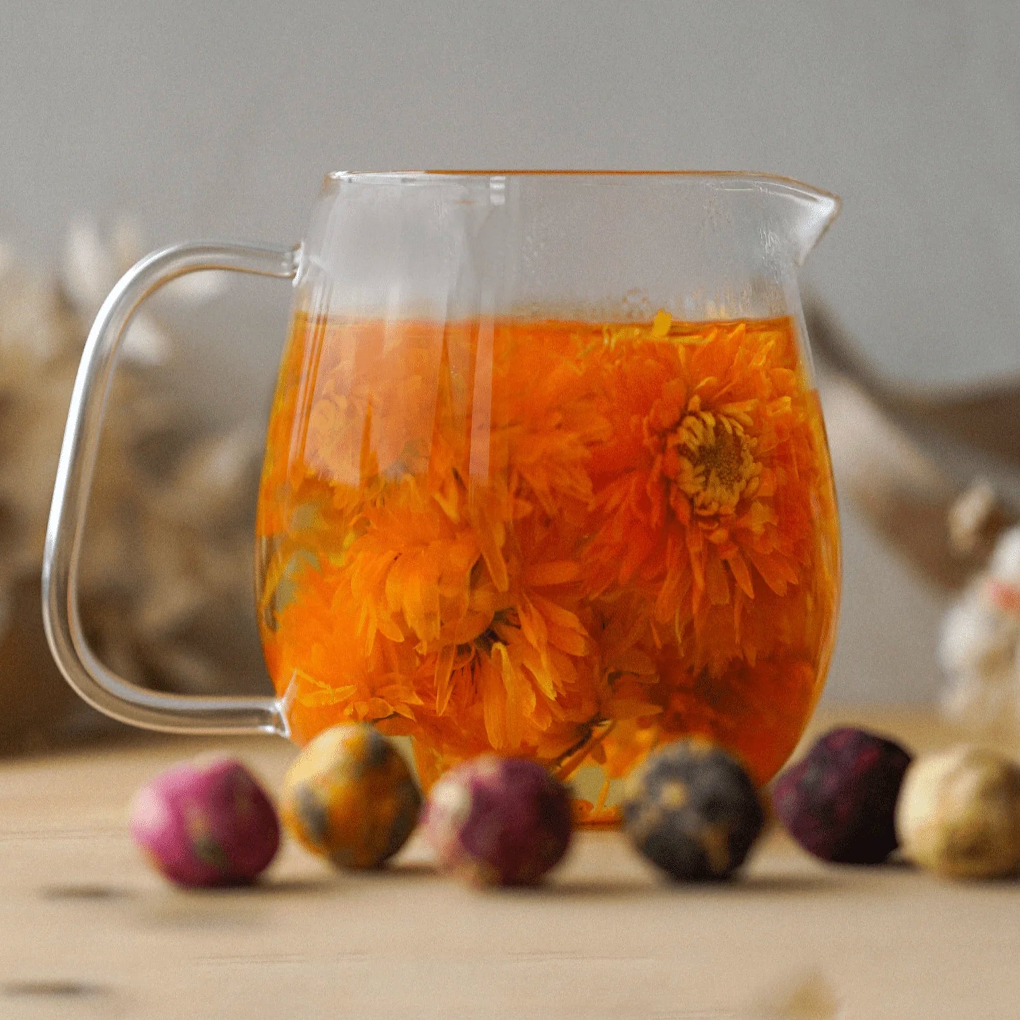 Glass pitcher filled with orange flower tea on a wooden surface with dried flowers.
