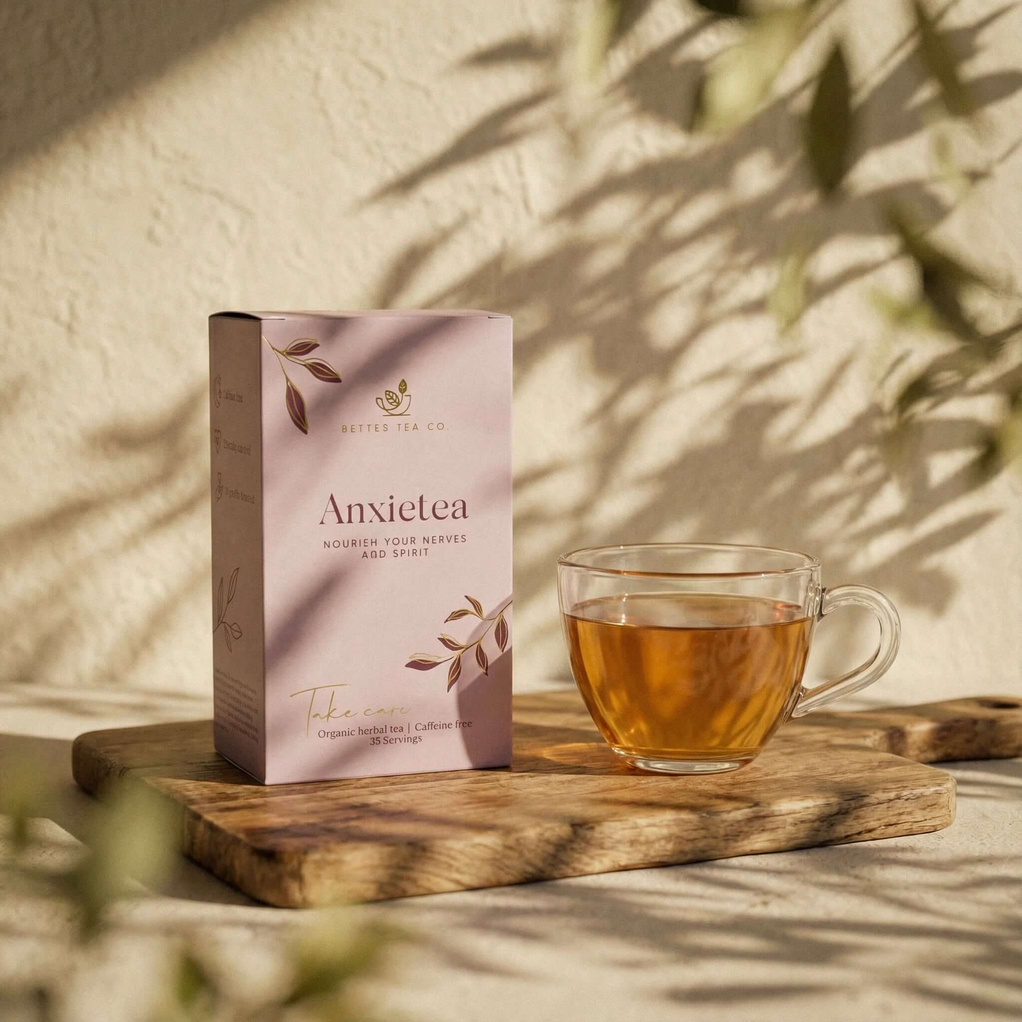 Better Tea Co. Wellness Tea - Anxietea herbal tea box beside glass cup of calming tea on wooden board in soft sunlight