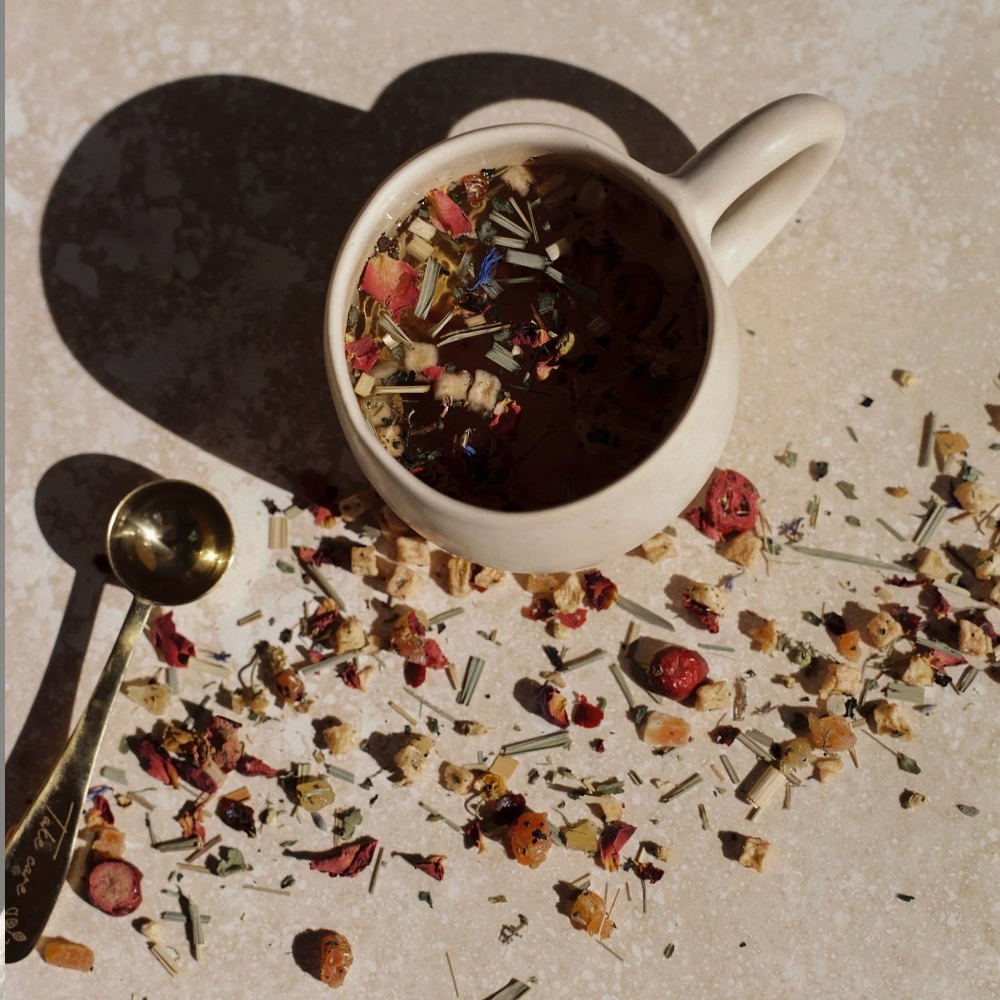 White mug filled with a dark liquid and dried herbs, surrounded by scattered herbs on a light surface.
