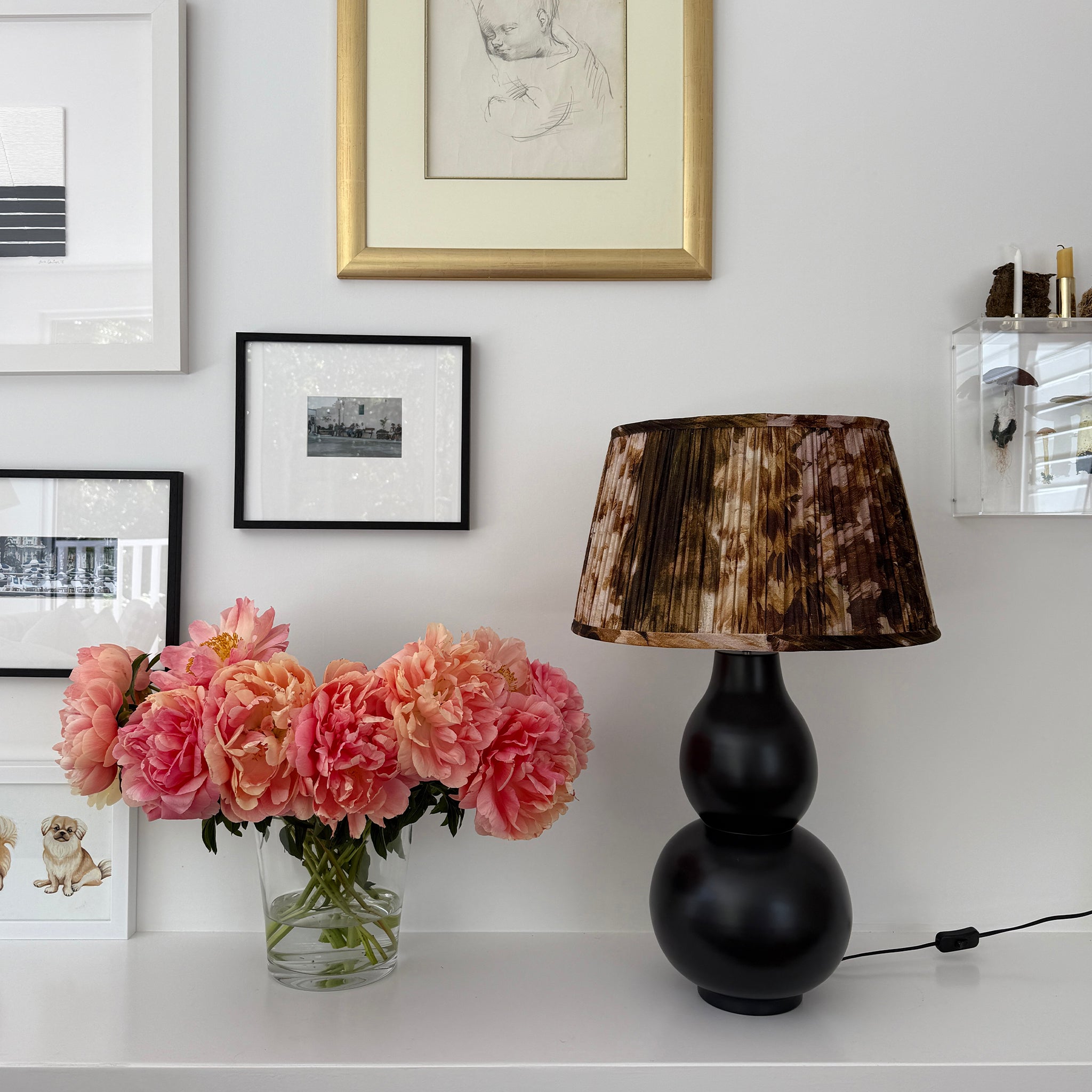 Black lamp with a pleated brown and black lampshade on a shelf with flowers.