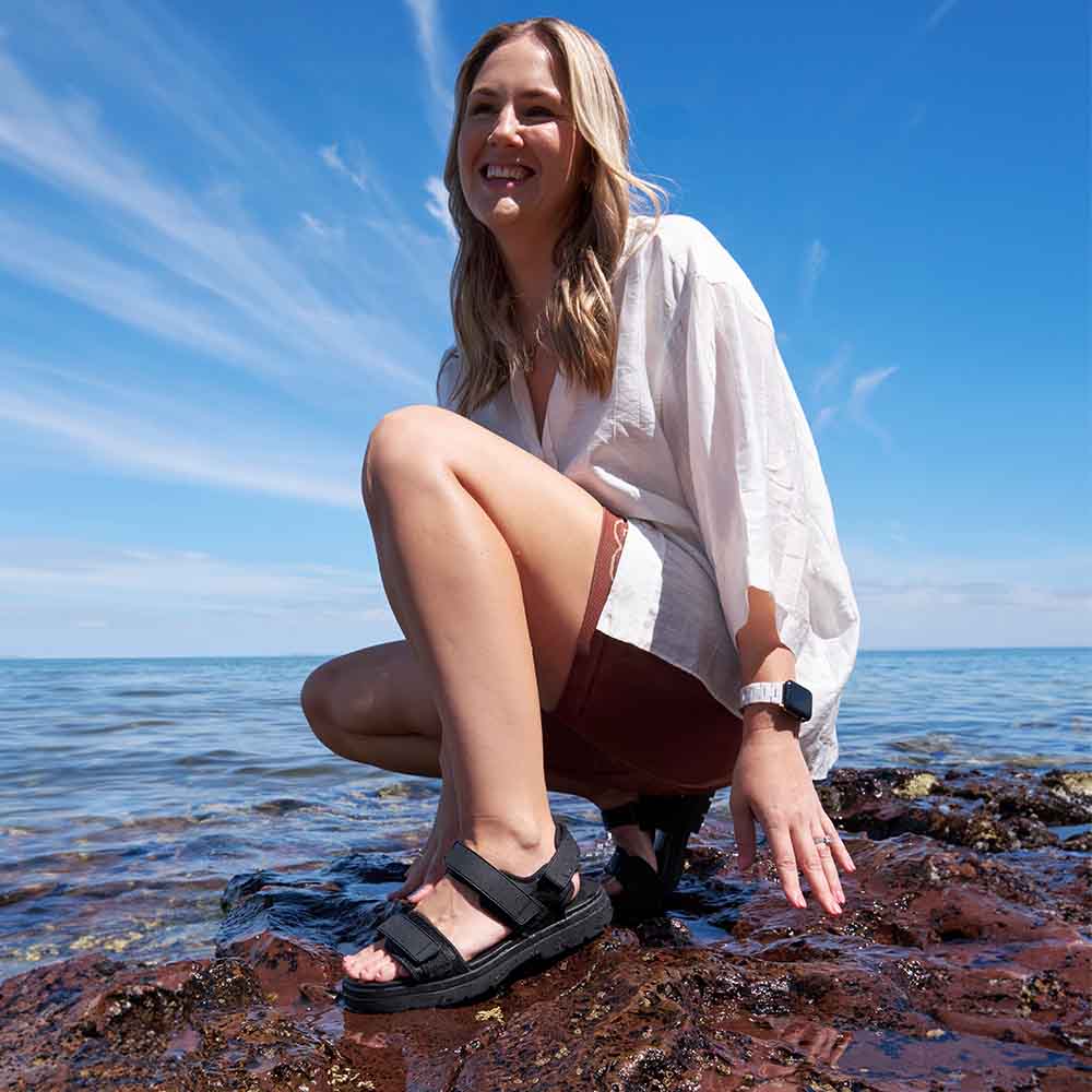 Woman squatting on rocks by the water wearing black sandals.