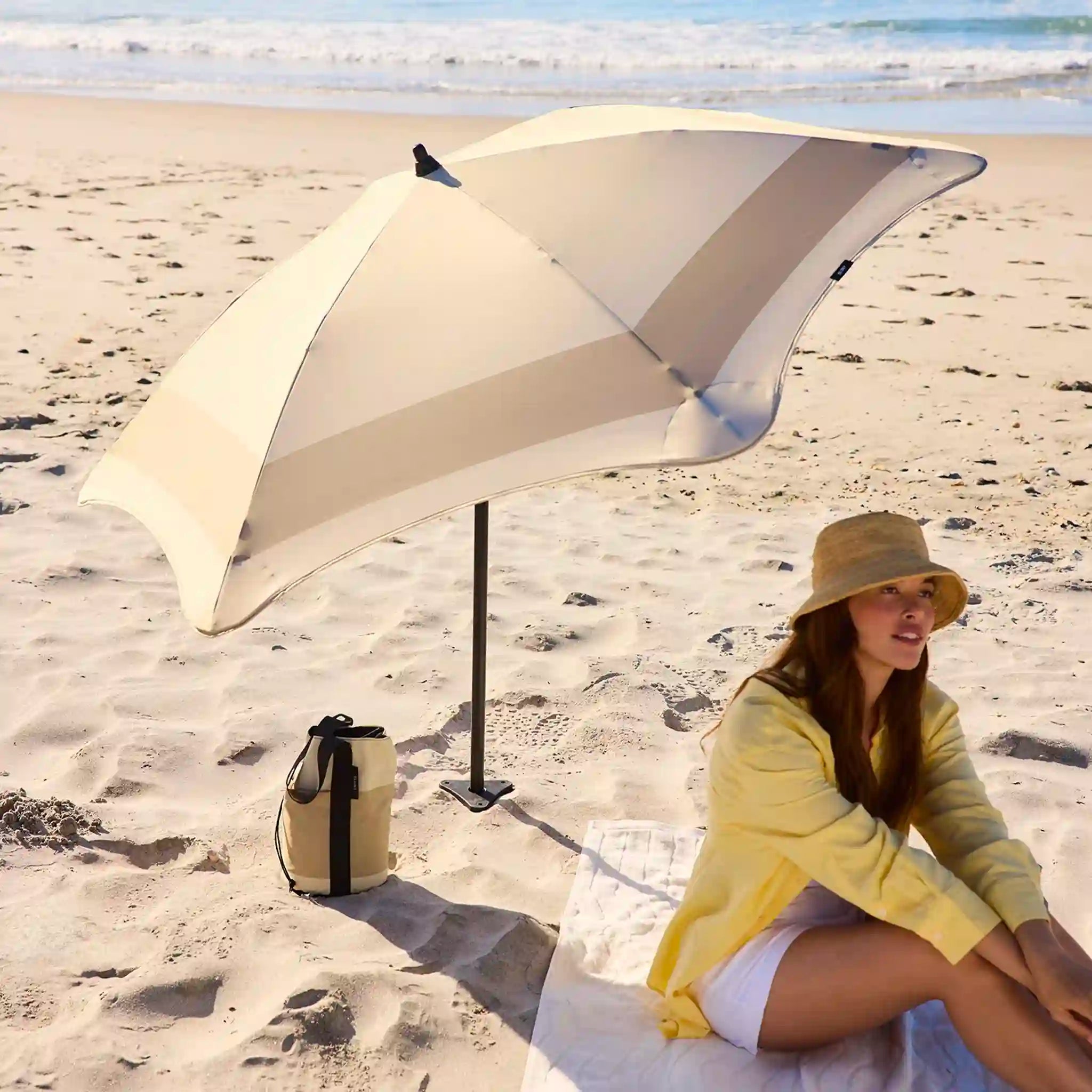 Woman sitting under a large beige beach umbrella on a sandy beach with ocean in the background