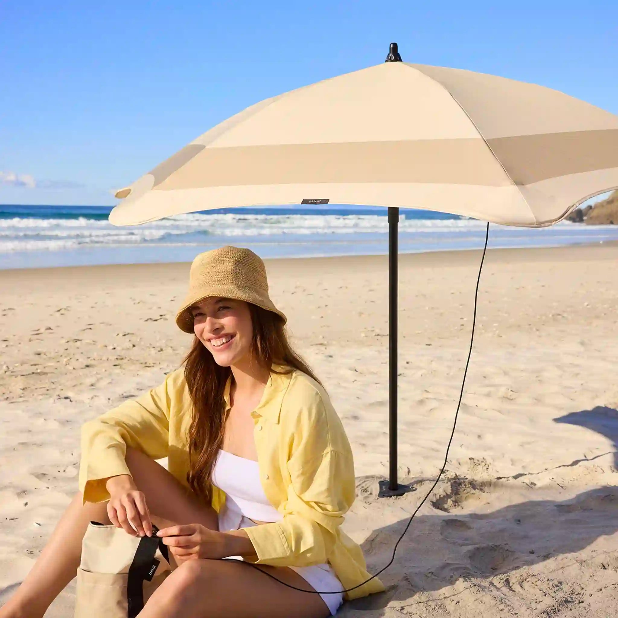 Woman sitting under a large beige beach umbrella on a sandy beach with ocean view.