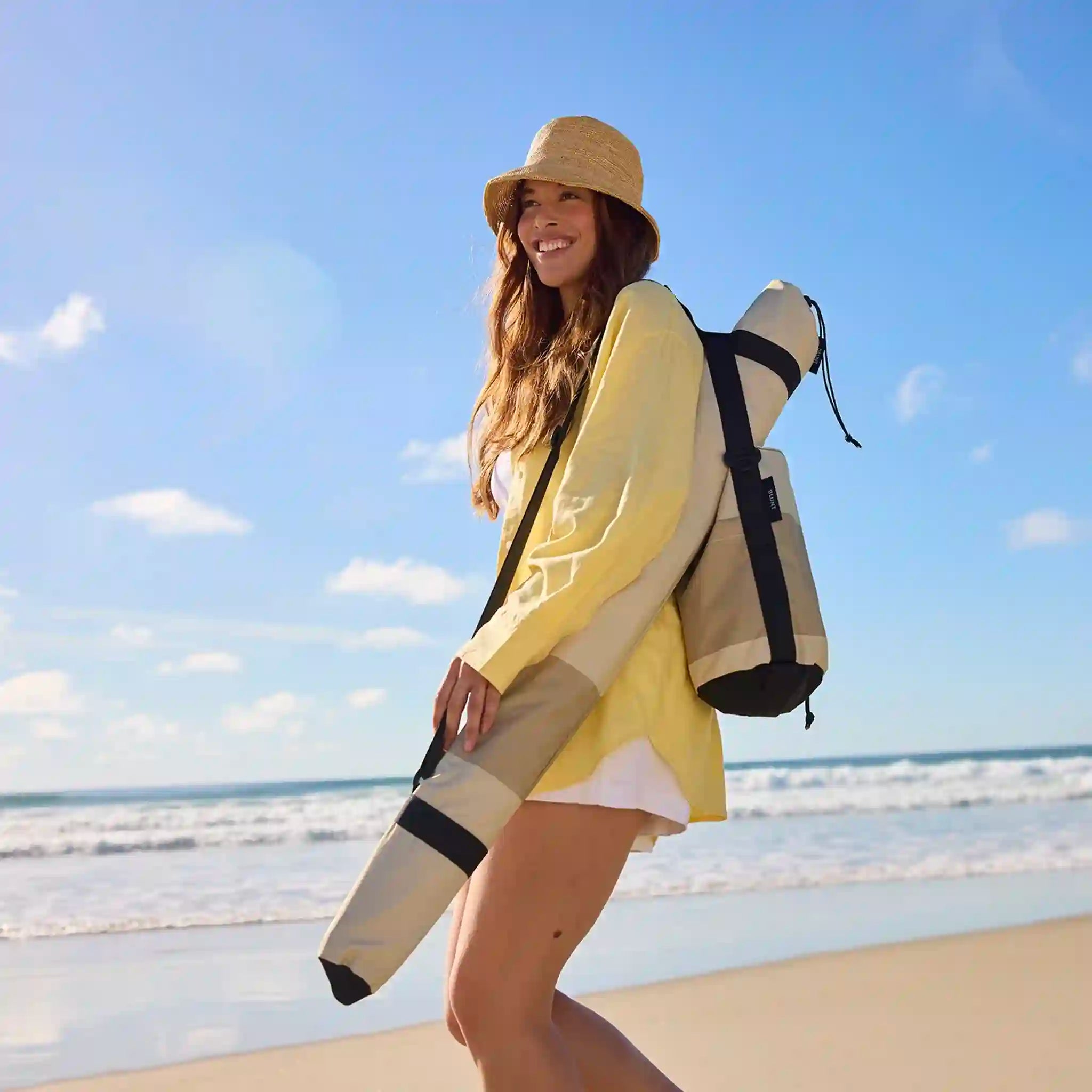 Woman on a beach holding a rolled-up mat with a blue sky and ocean background