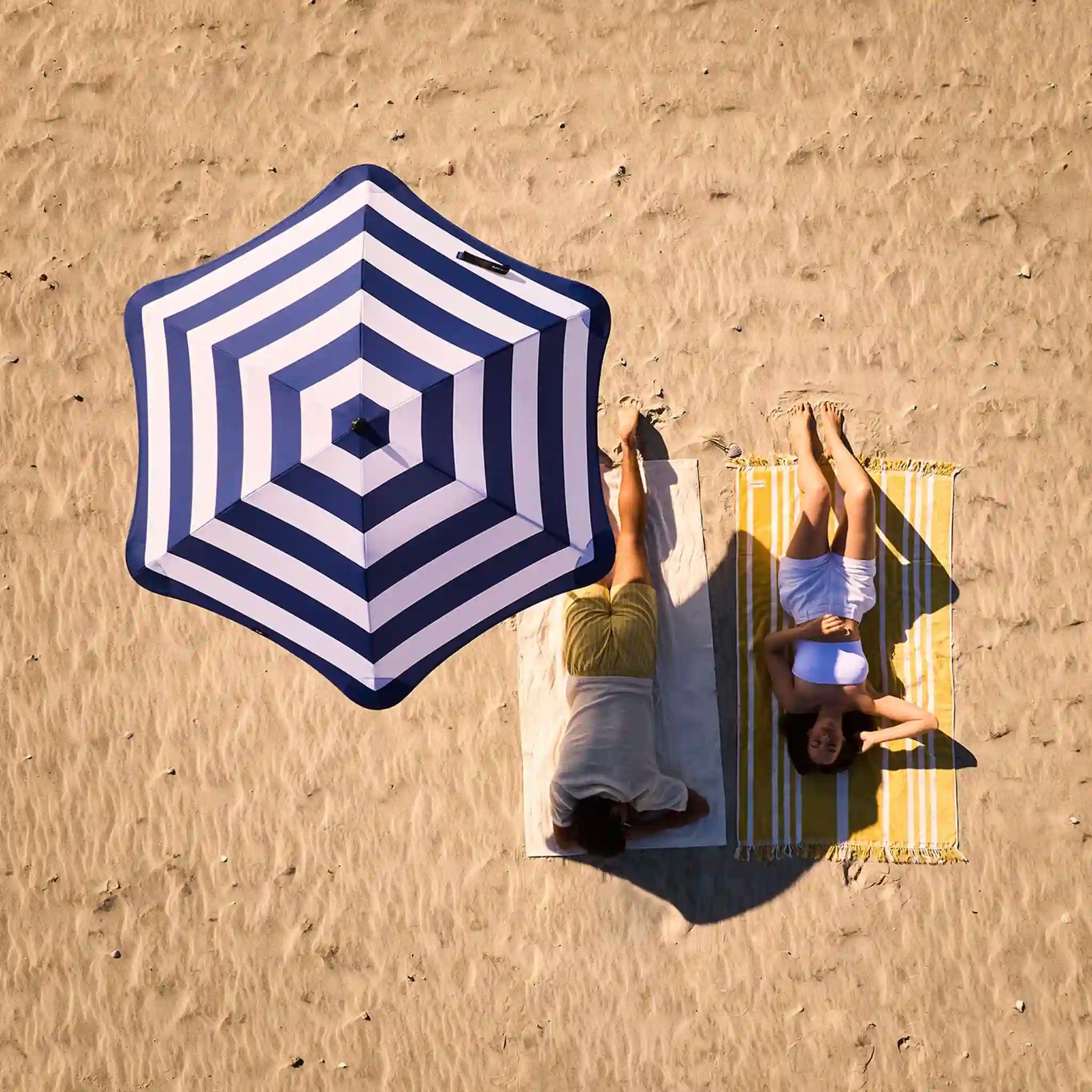 Two people lying on a beach with a large blue and white striped umbrella and matching beach towel.