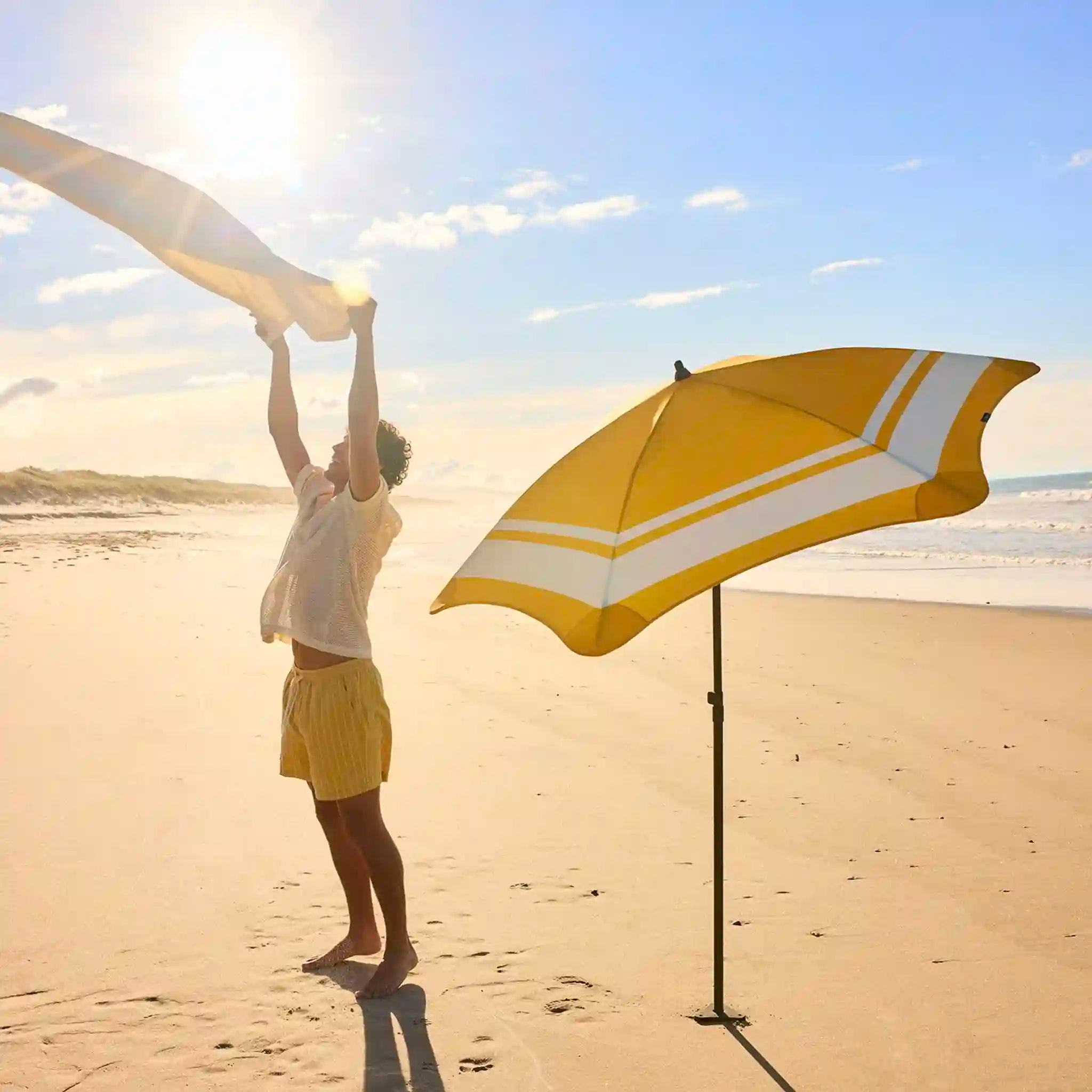 Person setting up a yellow and white beach umbrella on a sandy beach with ocean view.