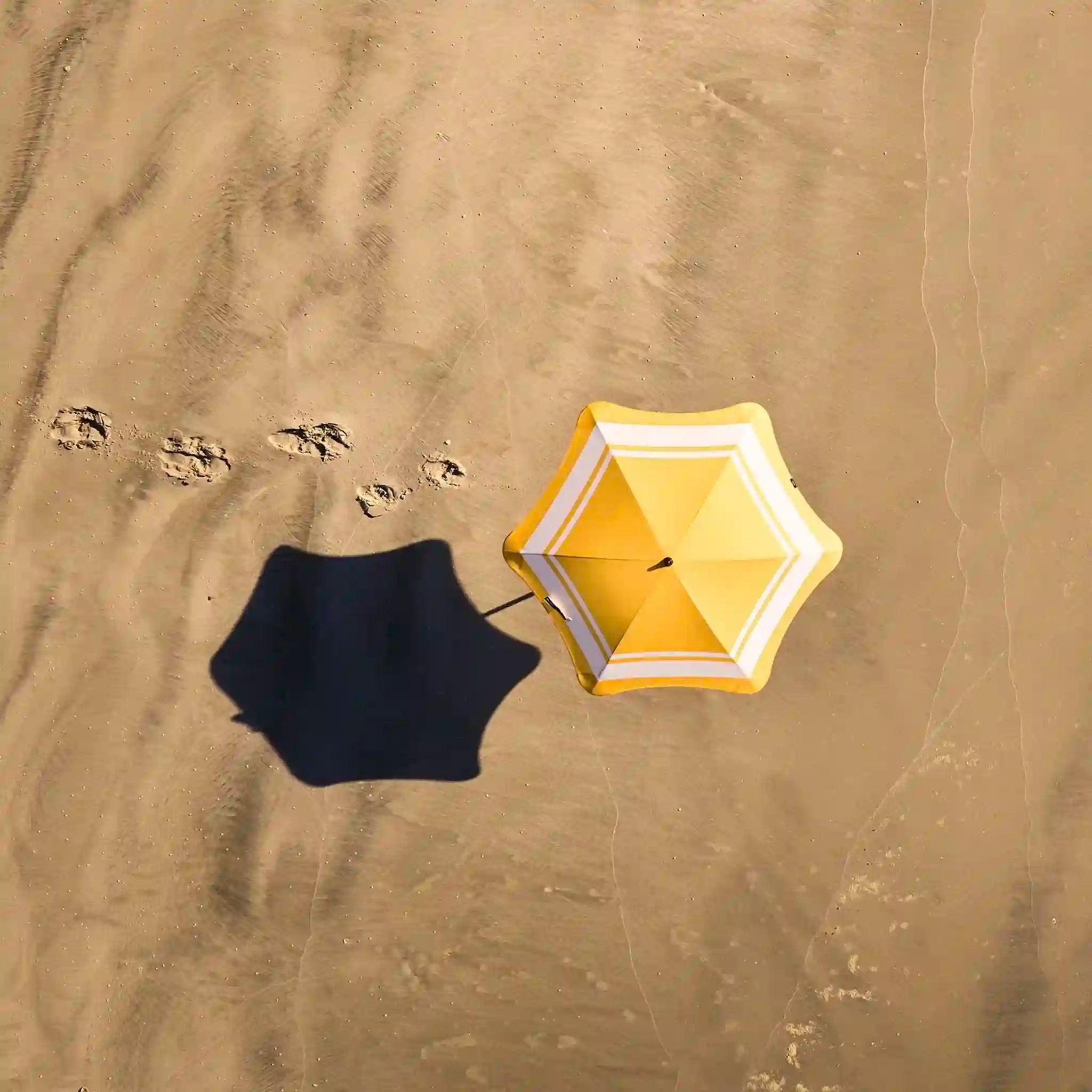 Yellow beach umbrella casting a shadow on sand with footprints nearby