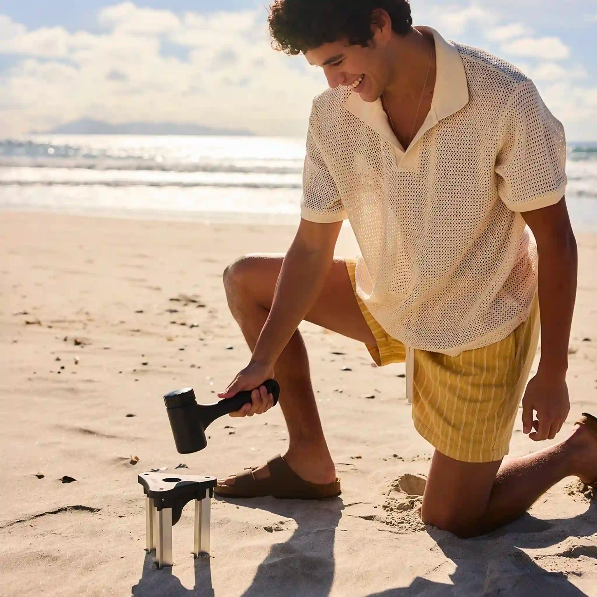 Man on a beach holding a mallet over a beach umbrella holder.