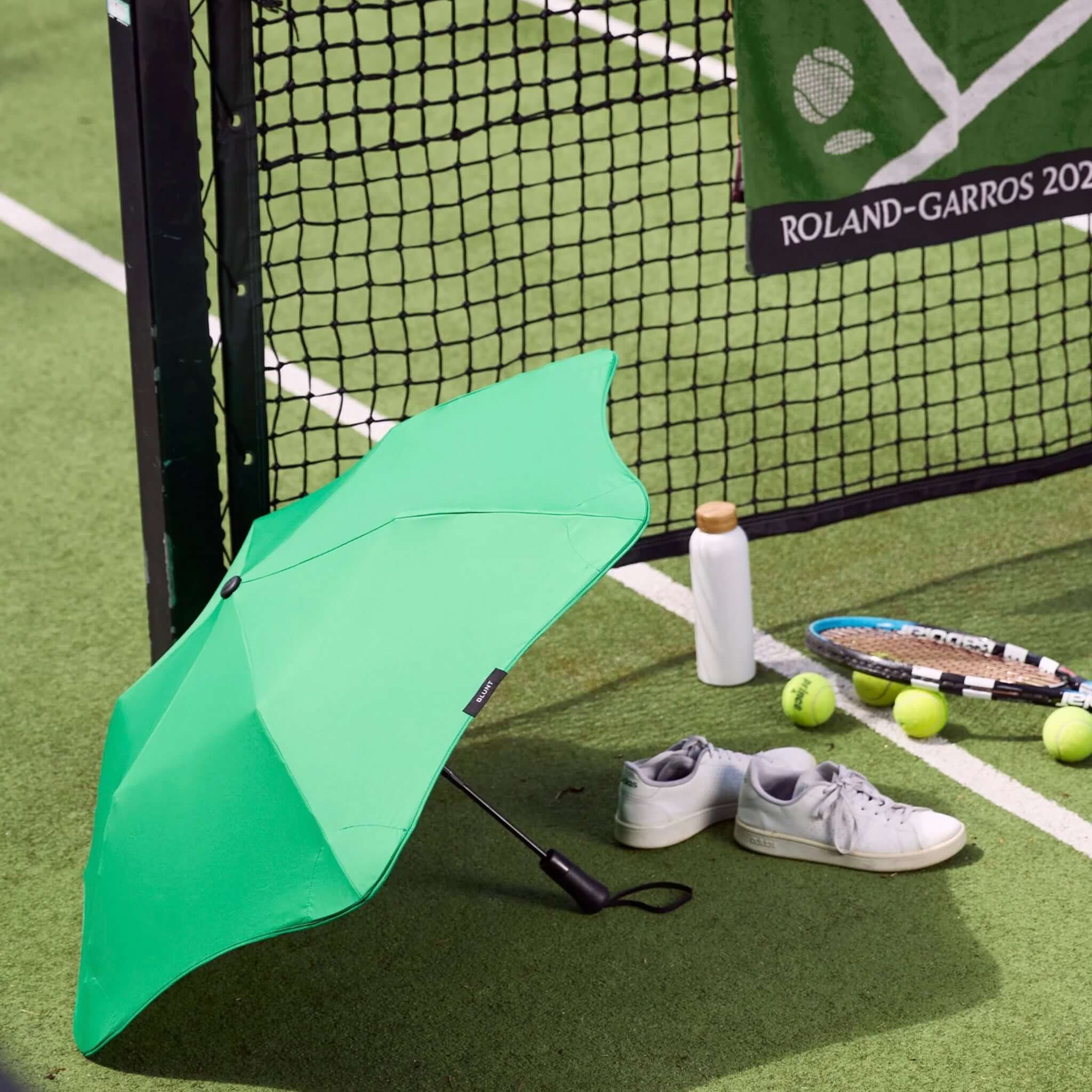 Green umbrella, white sneakers, water bottle, and tennis balls on a tennis court with Roland-Garros 2021 branding.