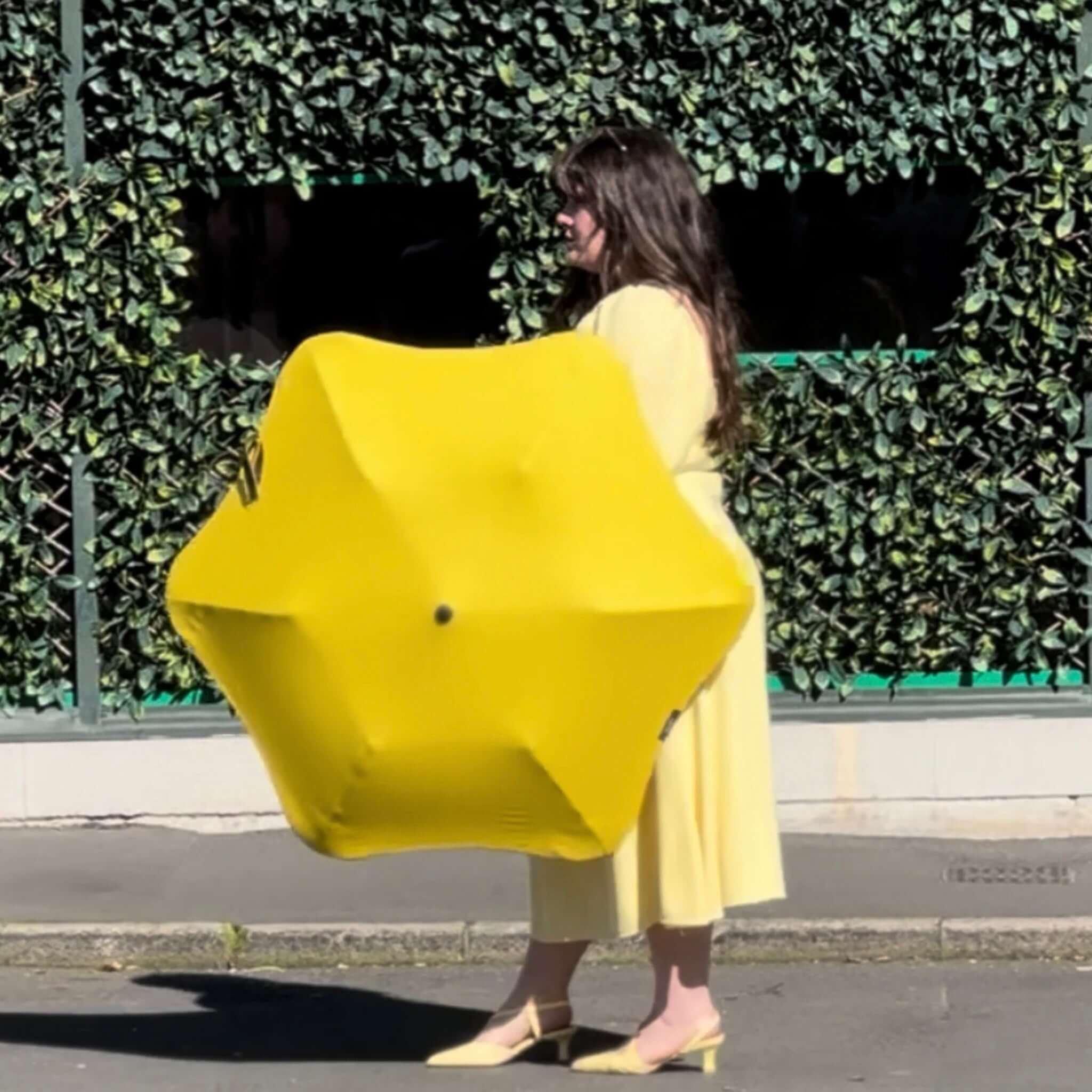 Person holding a large yellow umbrella against a green hedge background