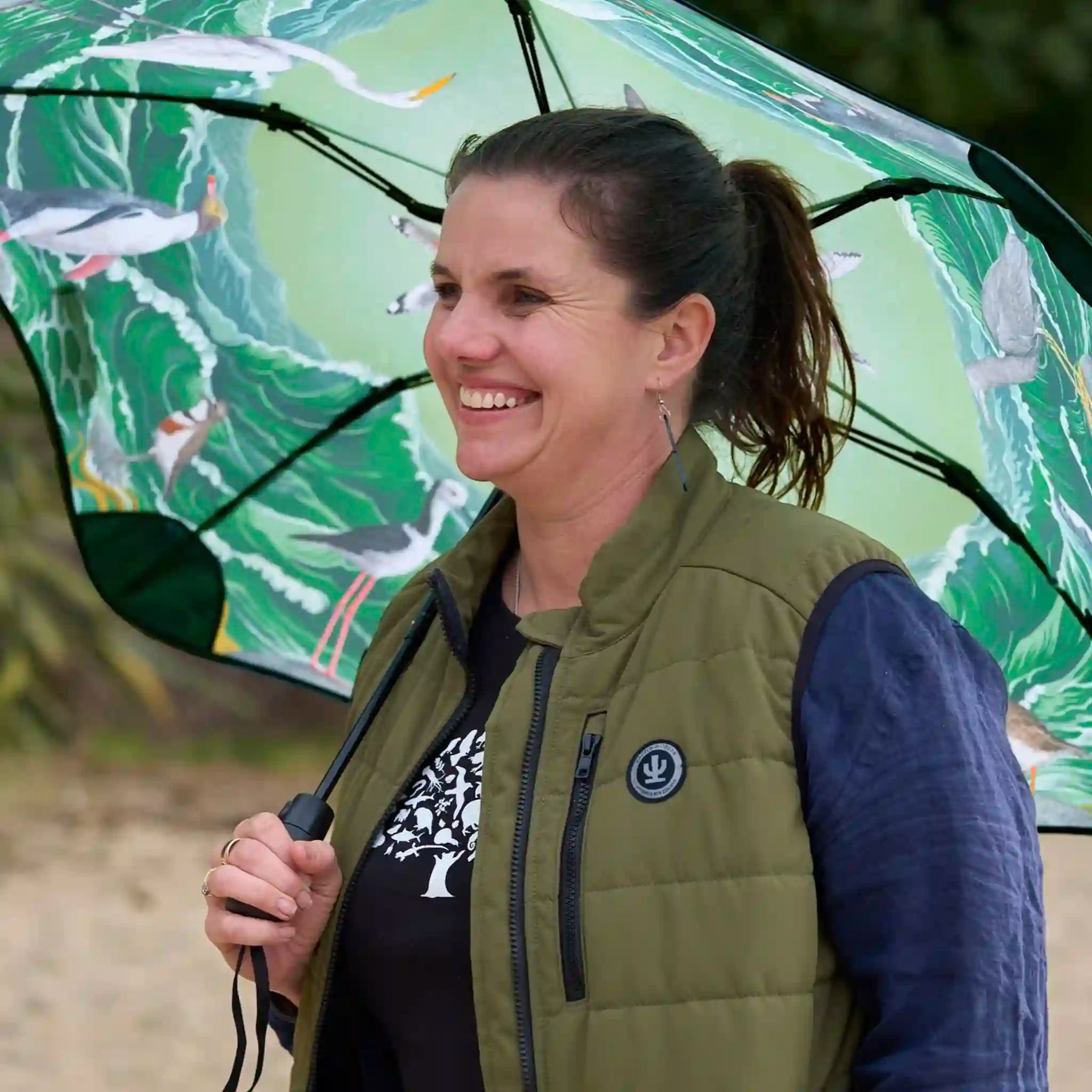 Woman holding a green umbrella with bird design, wearing a green vest and blue shirt.