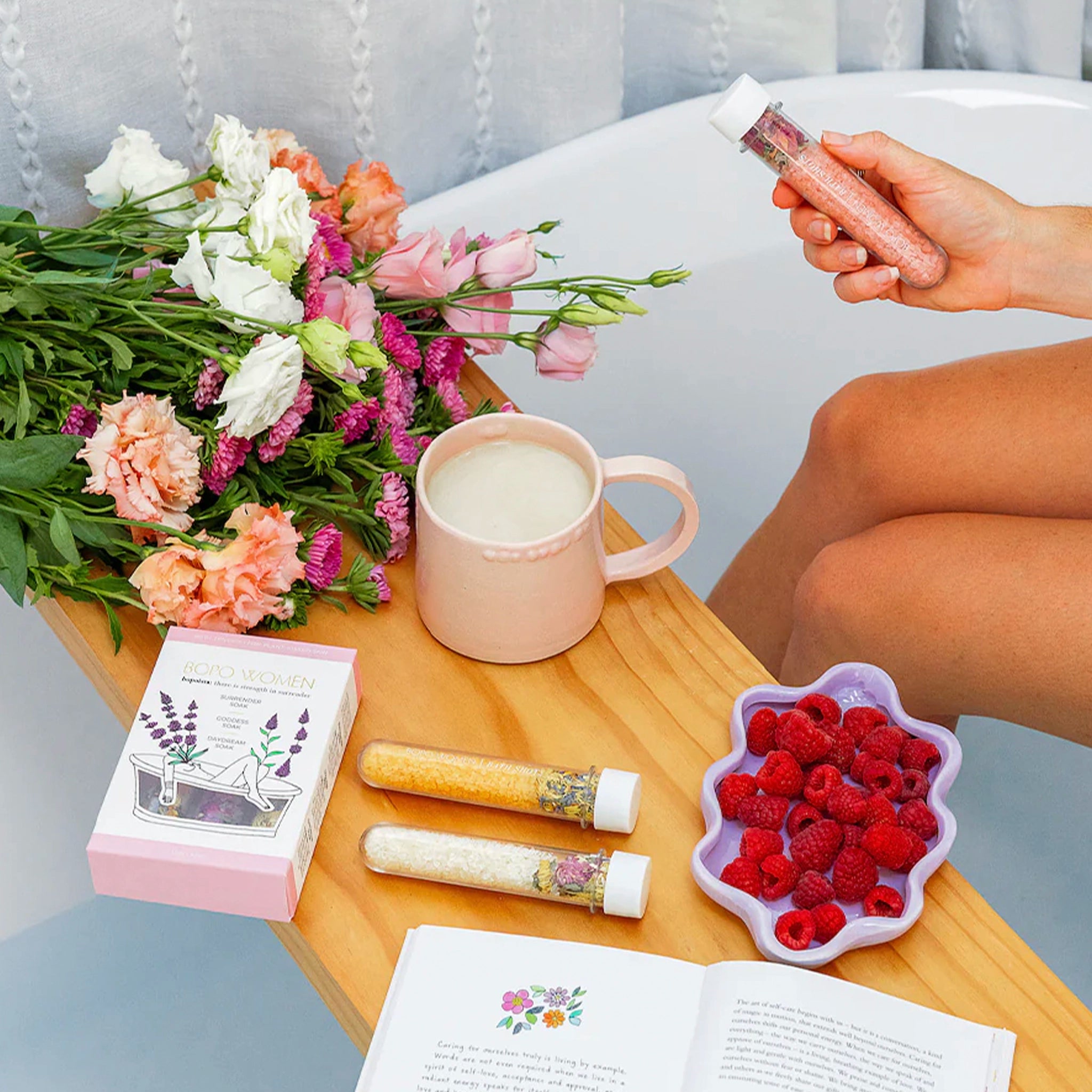 Person holding bath salts with a cup, flowers, and raspberries on a table