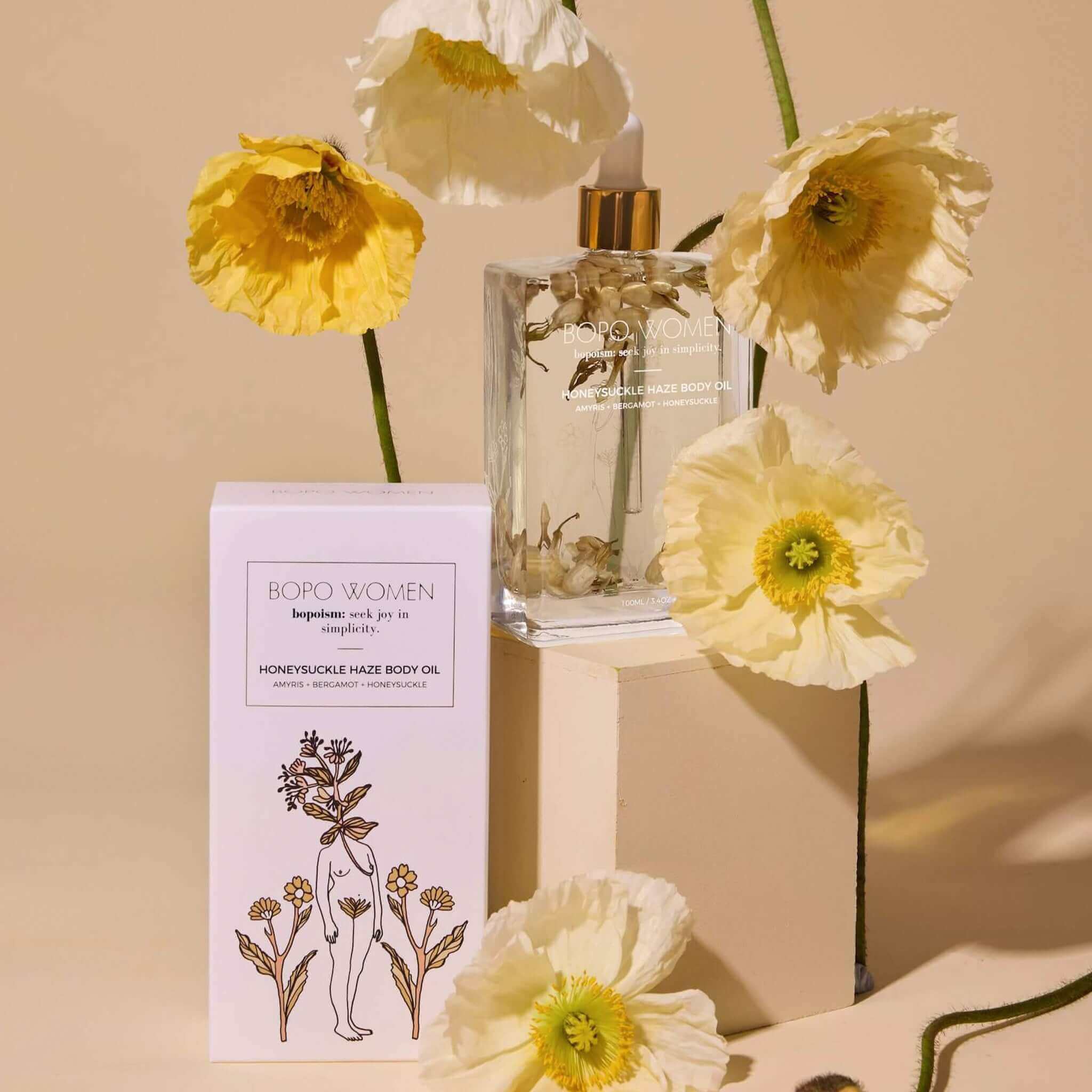 Bopo Women Body Oil - Honeysuckle Haze bottle and packaging surrounded by delicate flowers on a beige background.